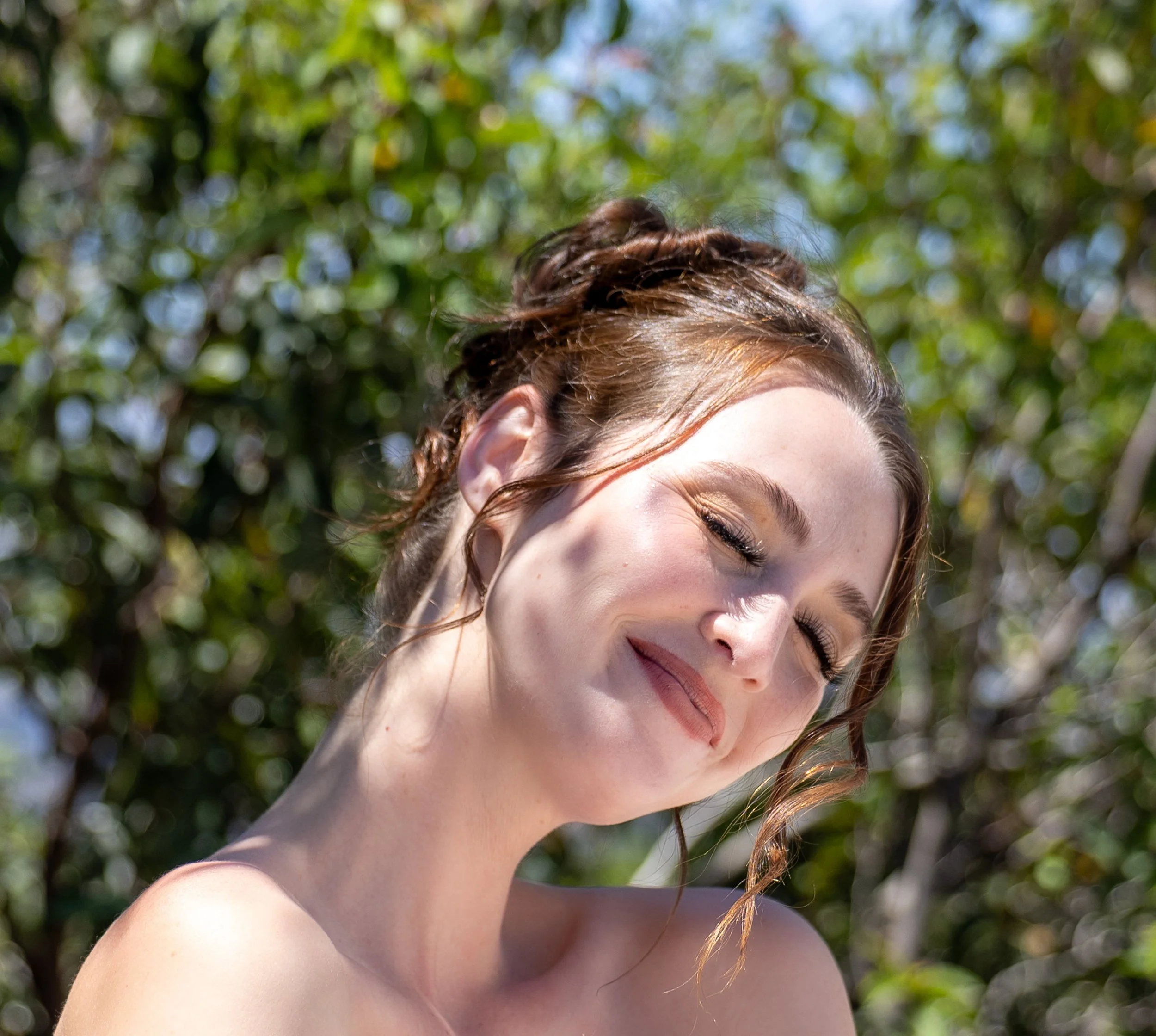 Close-up of a woman with red hair and closed eyes outdoors, with blurred green leaves in the background.