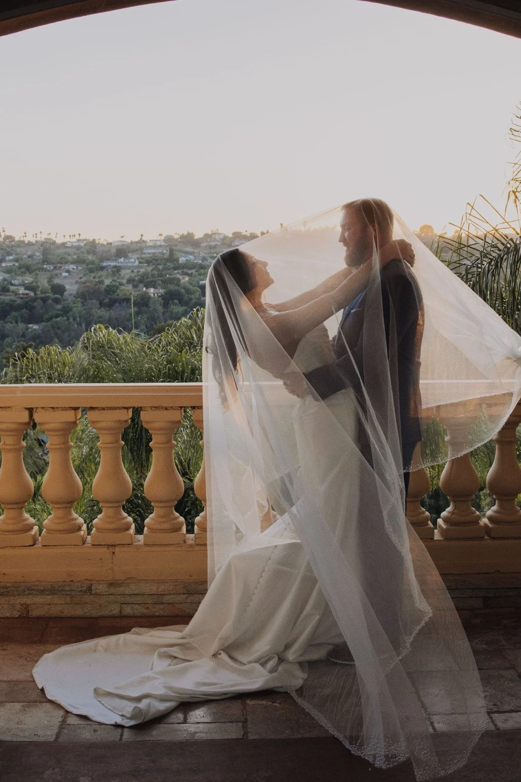 A bride and groom stand on a balcony, with the bride wrapping her veil around the groom, against a scenic sunset view of hills and trees.