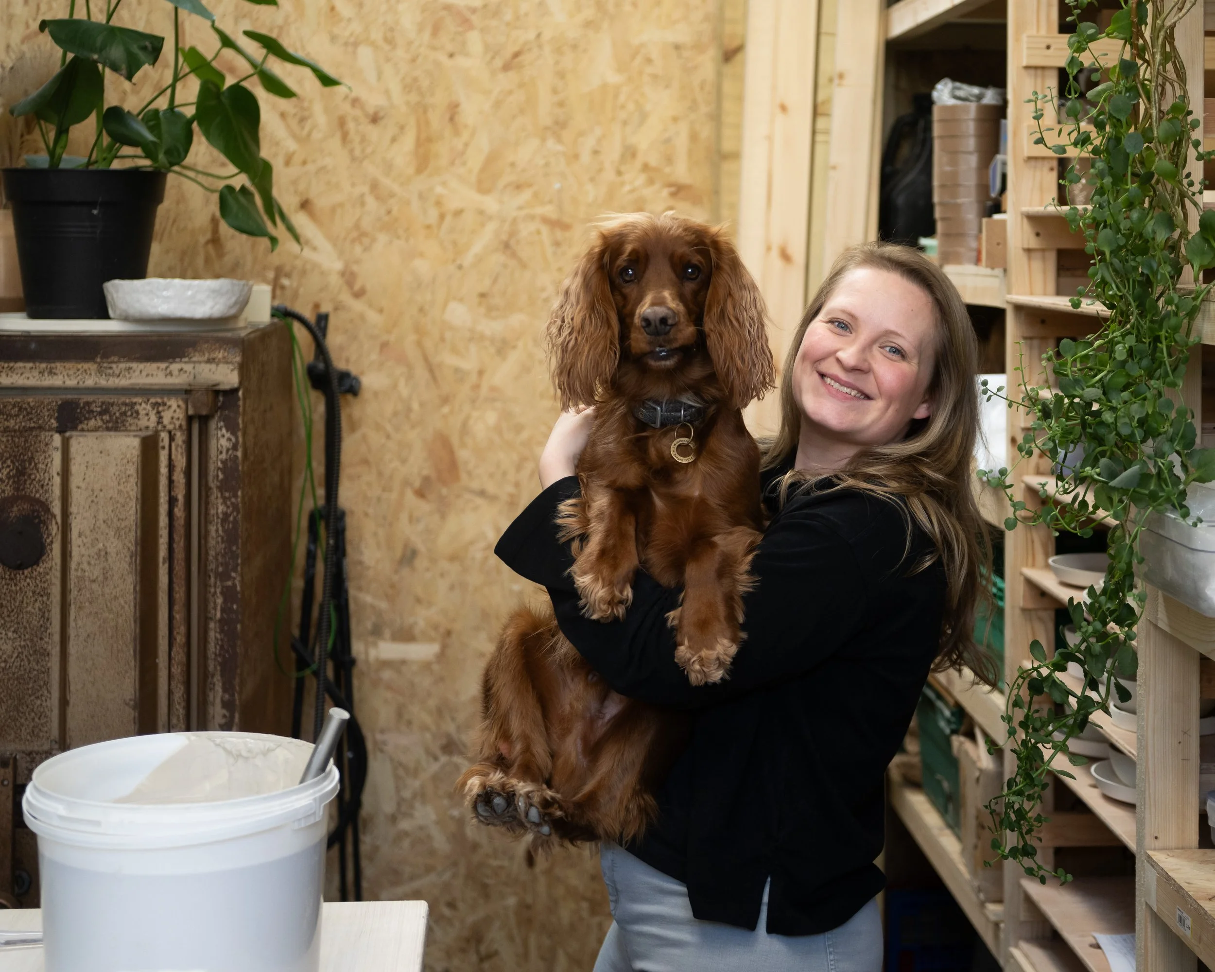 A woman holding a small brown dog with long ears in a woodworking studio. The dog is facing forward and the woman is smiling. There are potted plants, wooden shelves, and a container of white material on the table.