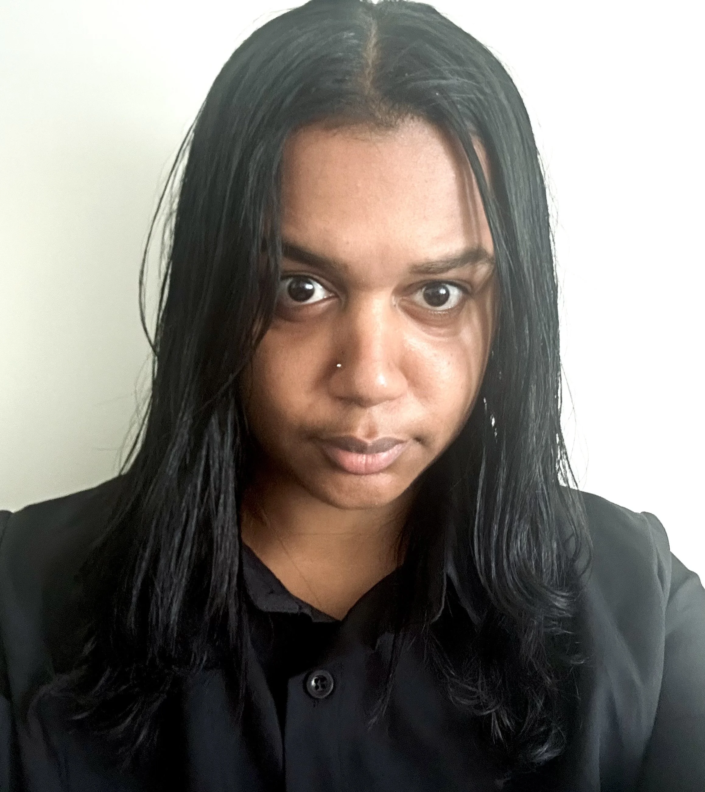 Close-up of a woman with long black hair, a nose piercing, and wearing a black shirt, standing against a plain white wall.