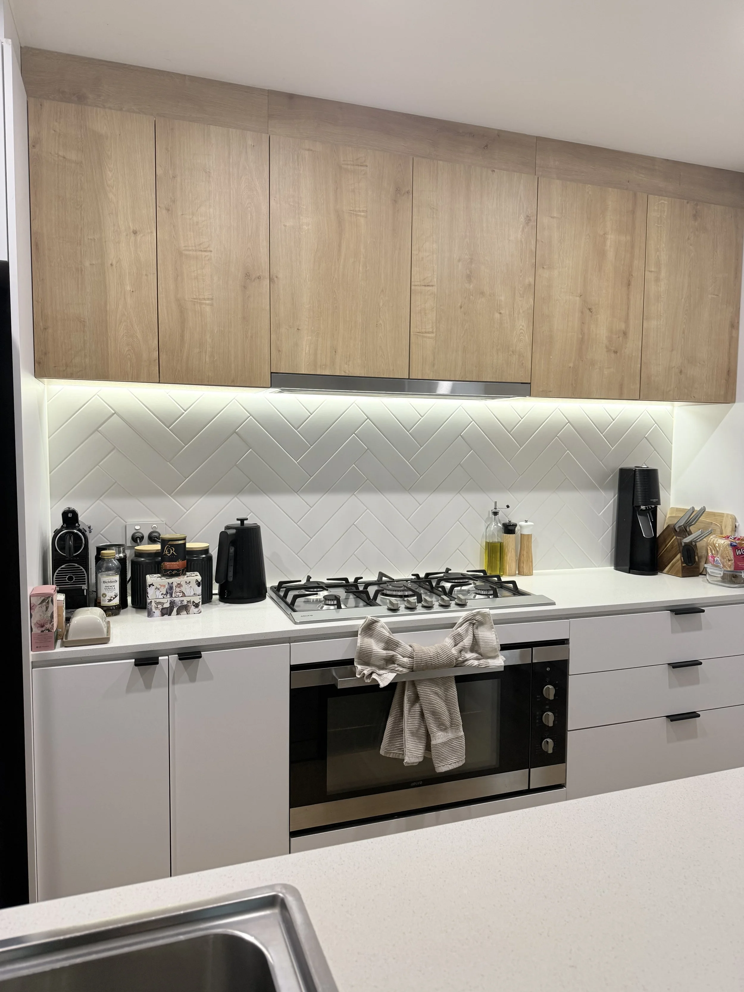 Modern kitchen with white cabinetry, wood upper cabinets, and a white tile backsplash in a herringbone pattern. Items on the counter include a black coffee maker, jars, bottles of oil and vinegar, paper towels, and a dish drying rack. A beige and white striped dish towel hangs from the oven handle.