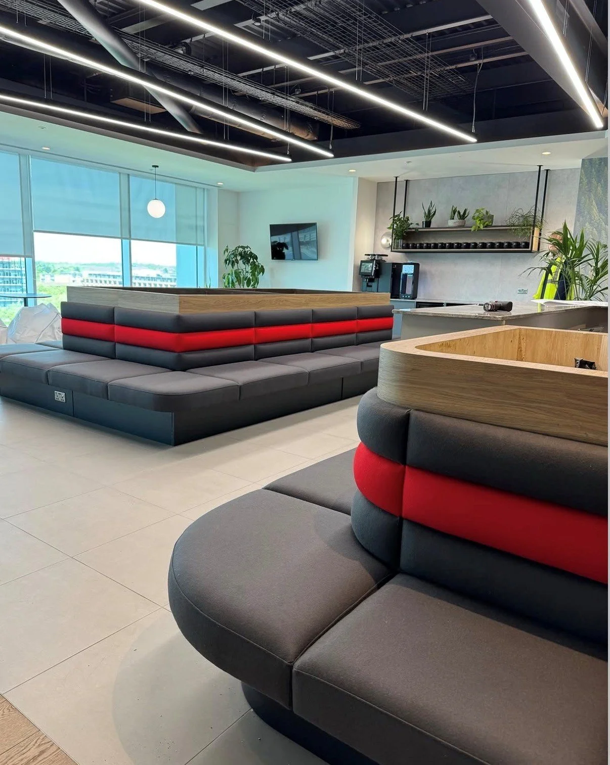 Modern lounge area with black and red cushioned seating, large windows, indoor plants, a wall-mounted TV, and a coffee station with coffee machines on a sleek countertop.