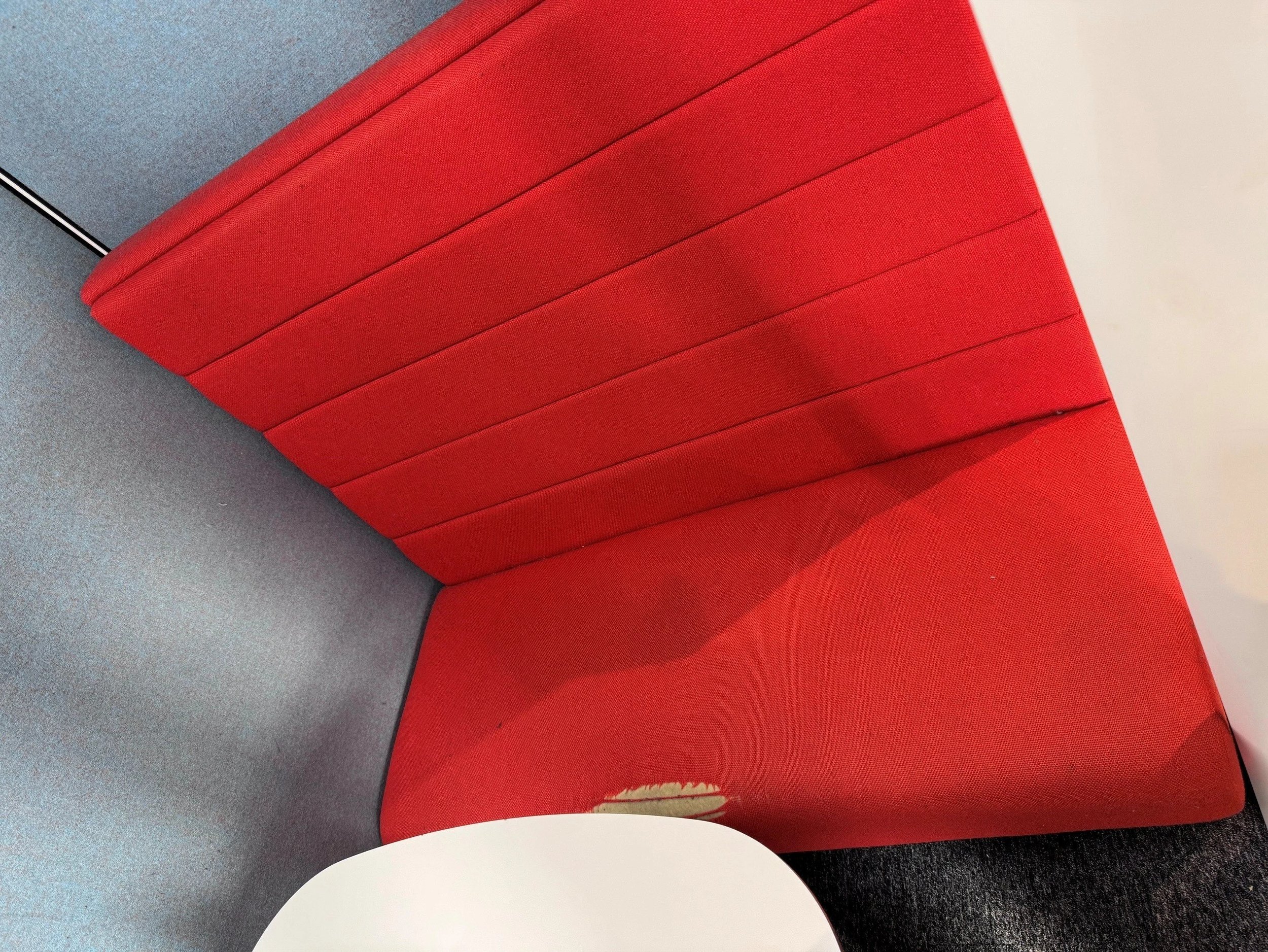 Red upholstered chair with visible wear at the bottom edge, sitting on a gray carpet next to a white surface or wall.