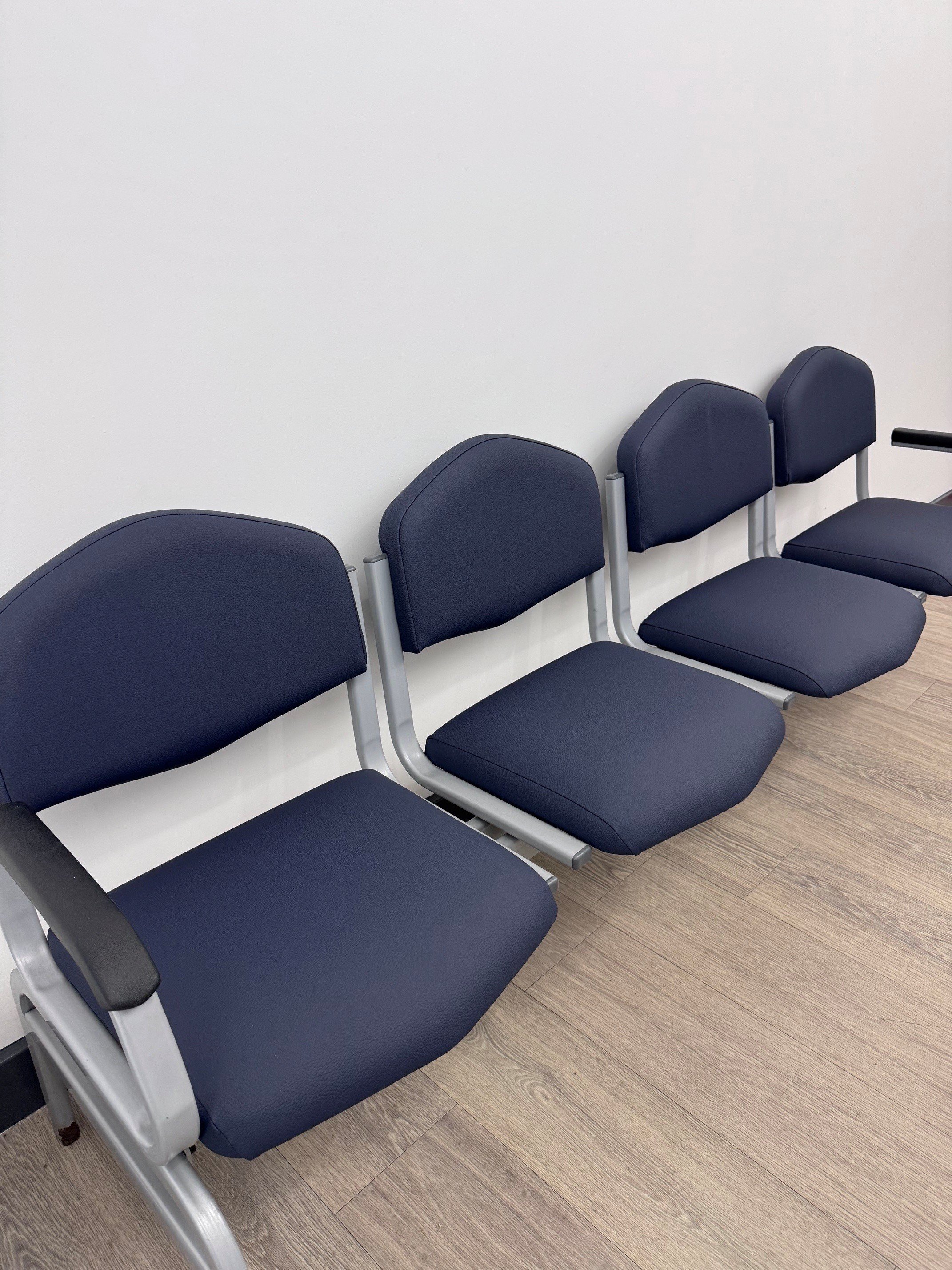 Row of blue cushioned chairs with metal frames against a plain white wall and wood flooring in a waiting area.