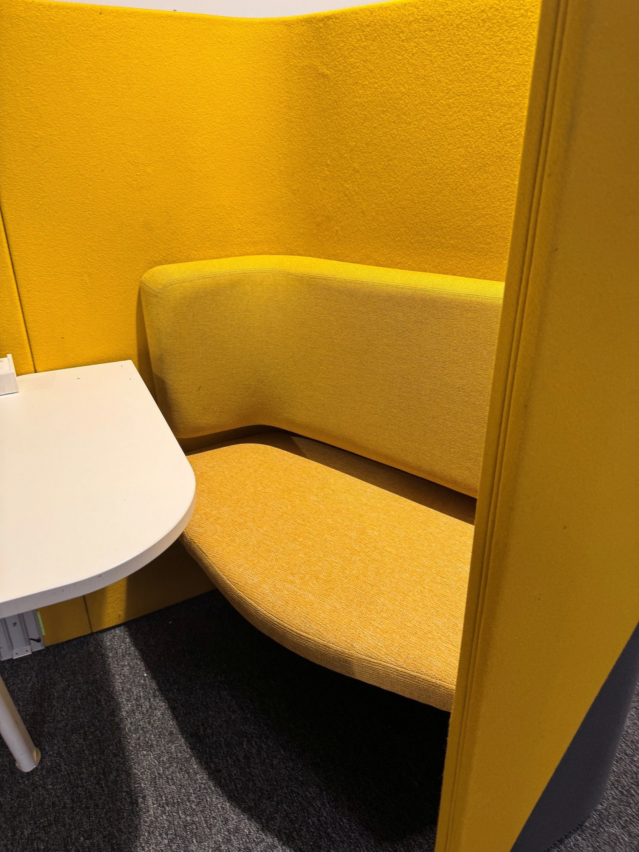 Yellow booth seating with a beige cushion, white table in a public or office space, and dark gray carpeted flooring.