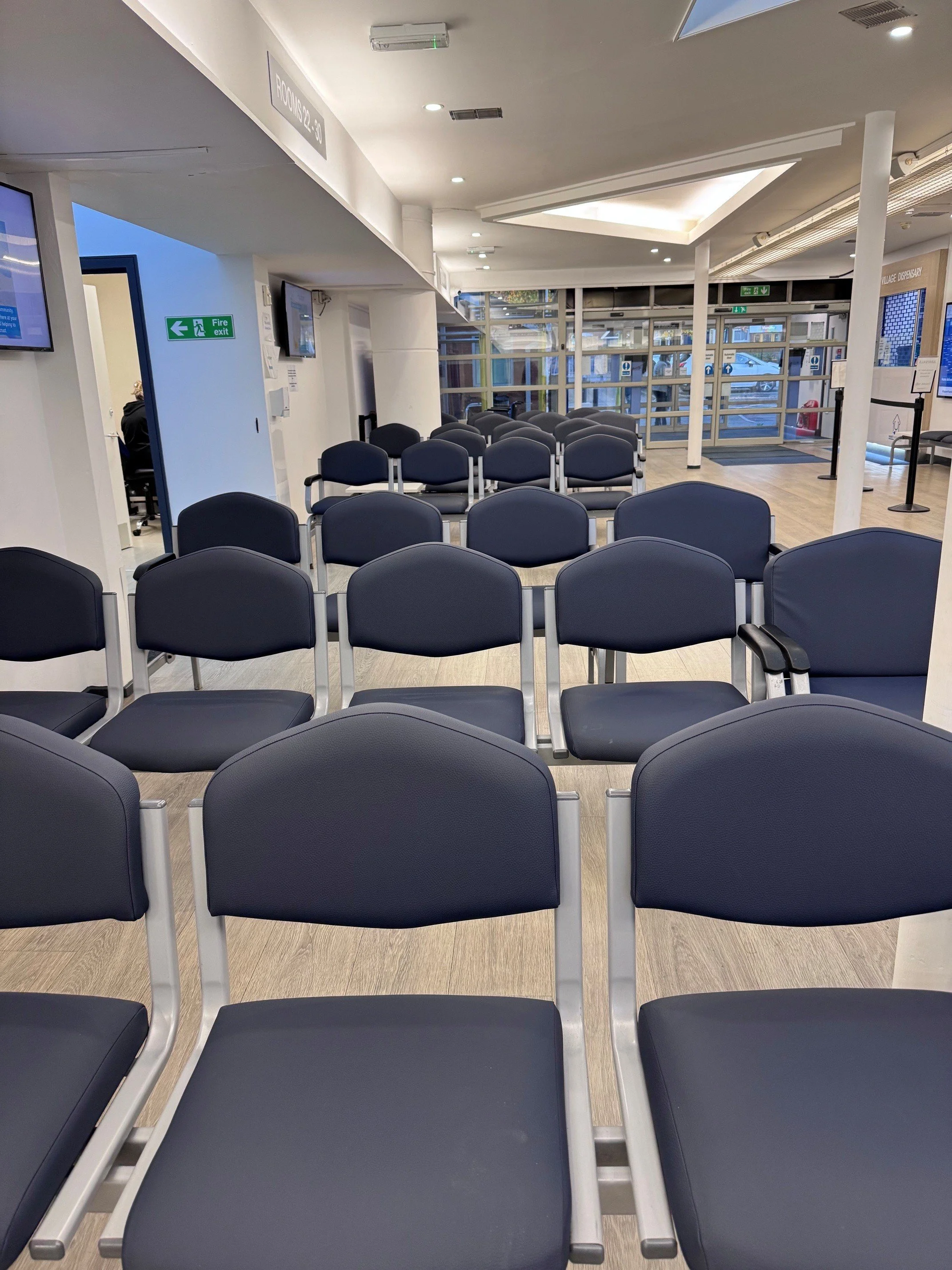 An empty airport waiting area with rows of black chairs, a fire exit sign, TV screens on the walls, and glass doors at the entrance.