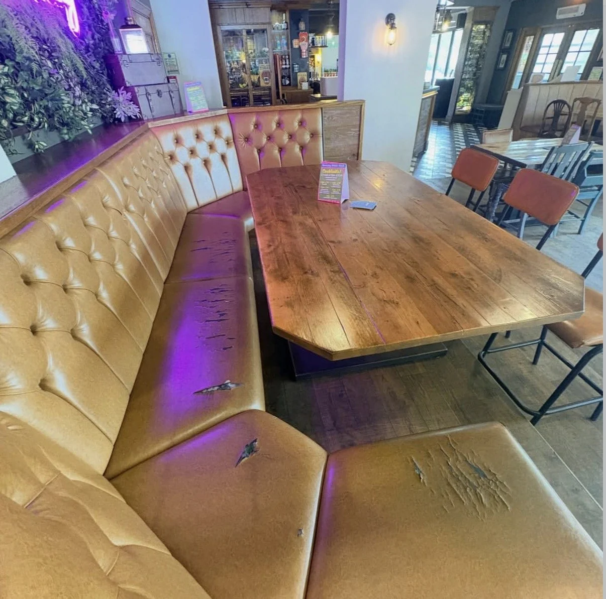A wooden table surrounded by a mustard yellow, tufted leather banquette with some torn and worn spots. In the background, a few dining chairs and tables are visible in a cozy restaurant or cafe setting with checkered and wooden flooring and large win