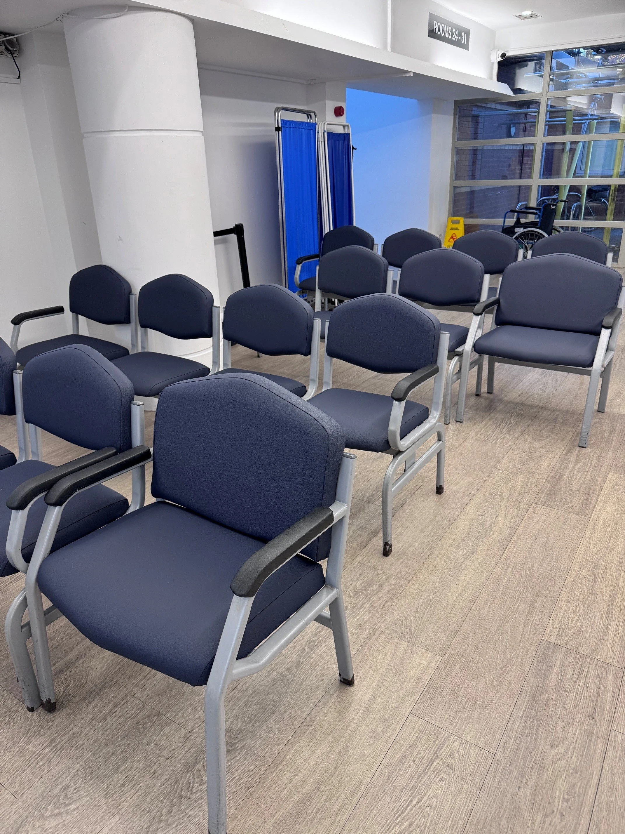 Empty waiting area with navy blue chairs arranged in rows, a blue foldable stretcher, a caution wet floor sign, and wheelchair near a large glass window at a medical facility.