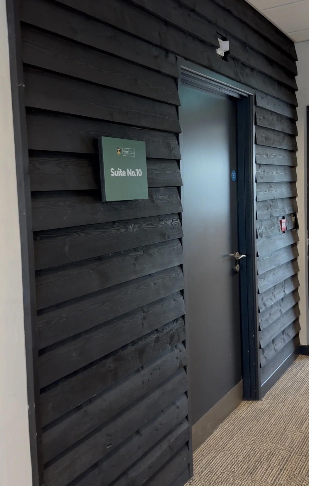 A modern office hallway with dark wooden panel walls, a gray door marked 'Suite No. 10', and a striped carpeted floor.