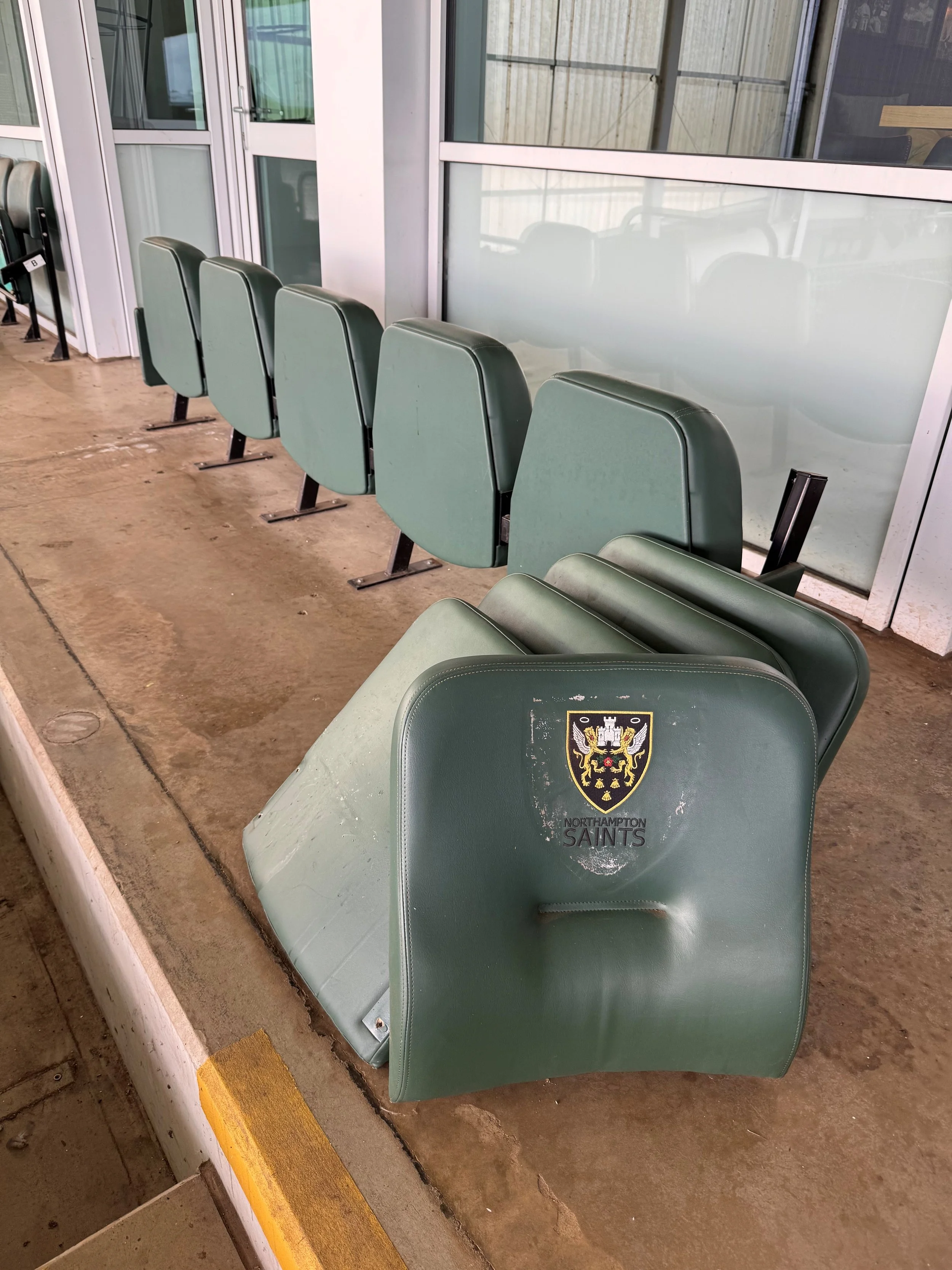 Empty row of green stadium seats with a Northhampton Saints logo on the backrest in the foreground, a concrete floor, and glass windows behind the seats.