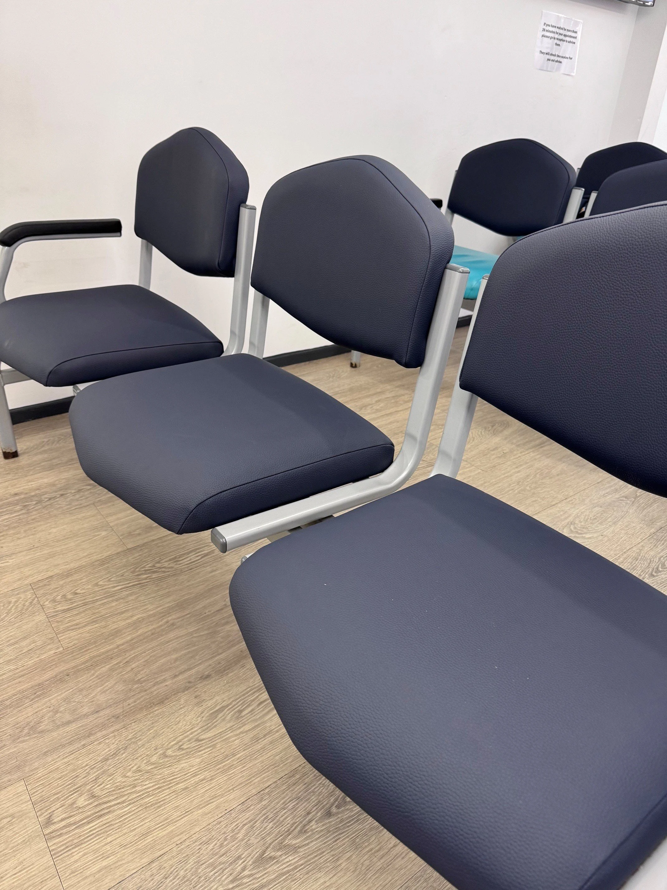 Multiple empty blue chairs in a waiting area with a white wall and a small sign in the background.