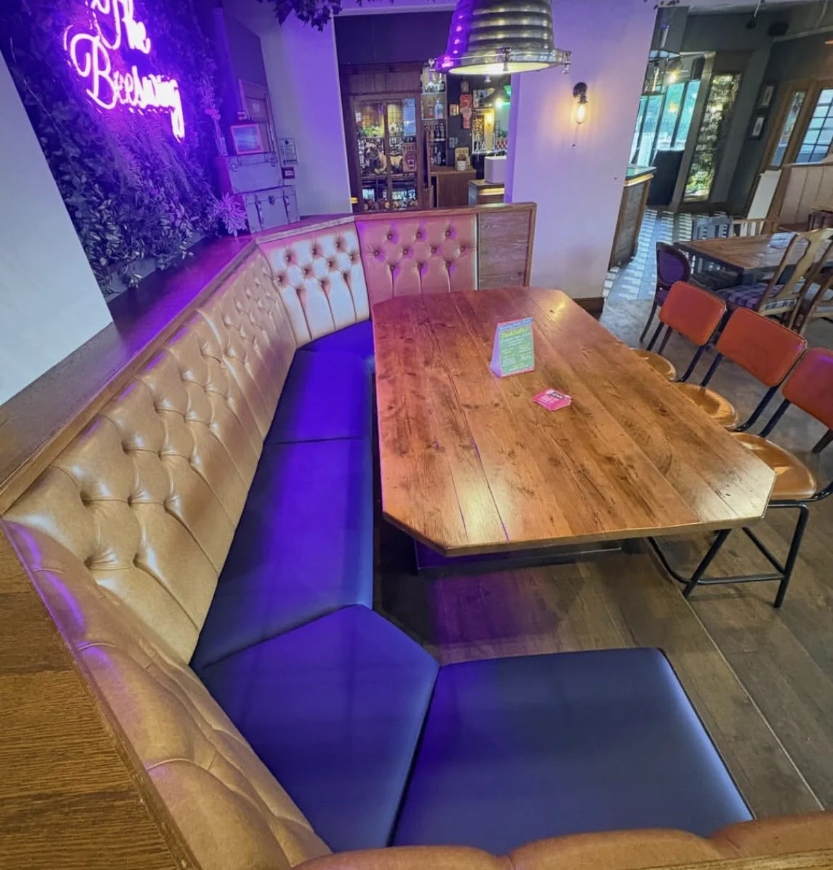 A cozy restaurant corner with a curved beige tufted leather banquette, a large wooden table, and orange chairs. Neon purple lighting reading 'The Beetroot' and colorful ambient lights create a vibrant atmosphere.