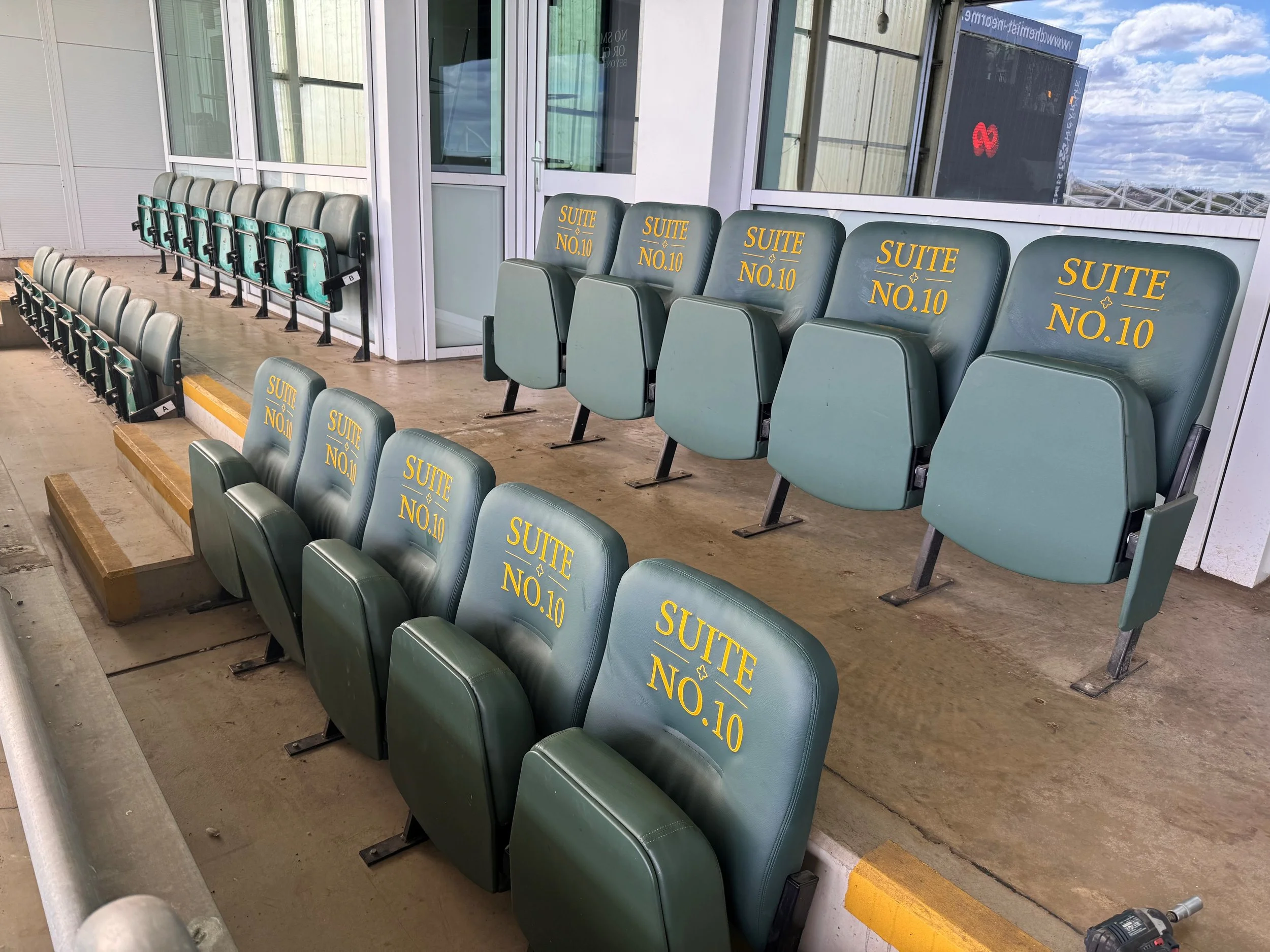 Multiple green seats on an outdoor airport terminal with 'SUITE NO.10' written in yellow on the backrests, positioned over a concrete floor with yellow safety line and glass windows behind.