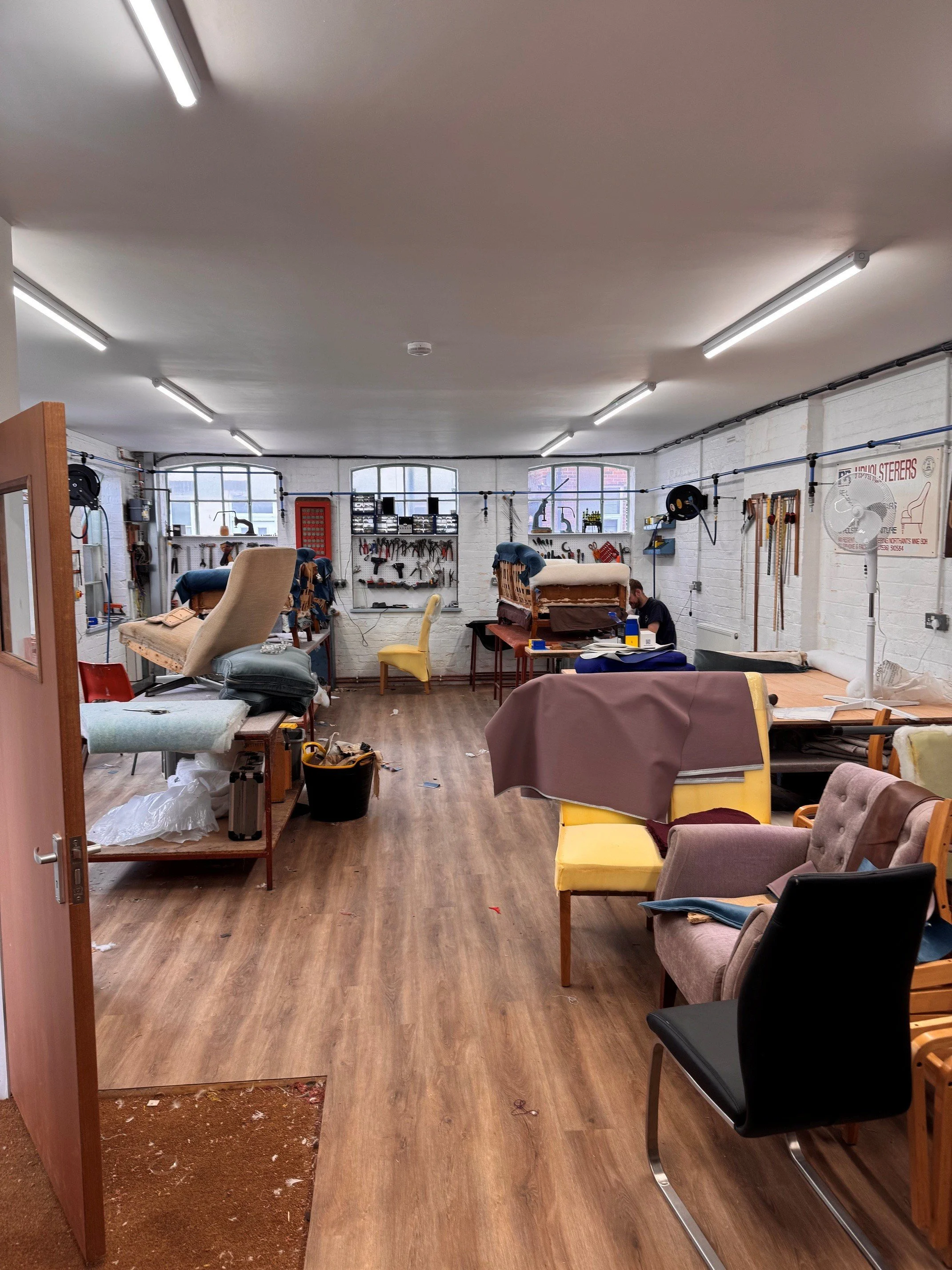 A cluttered workshop or craft room with various chairs, tools, and objects scattered around, and a white brick wall with windows in the background.