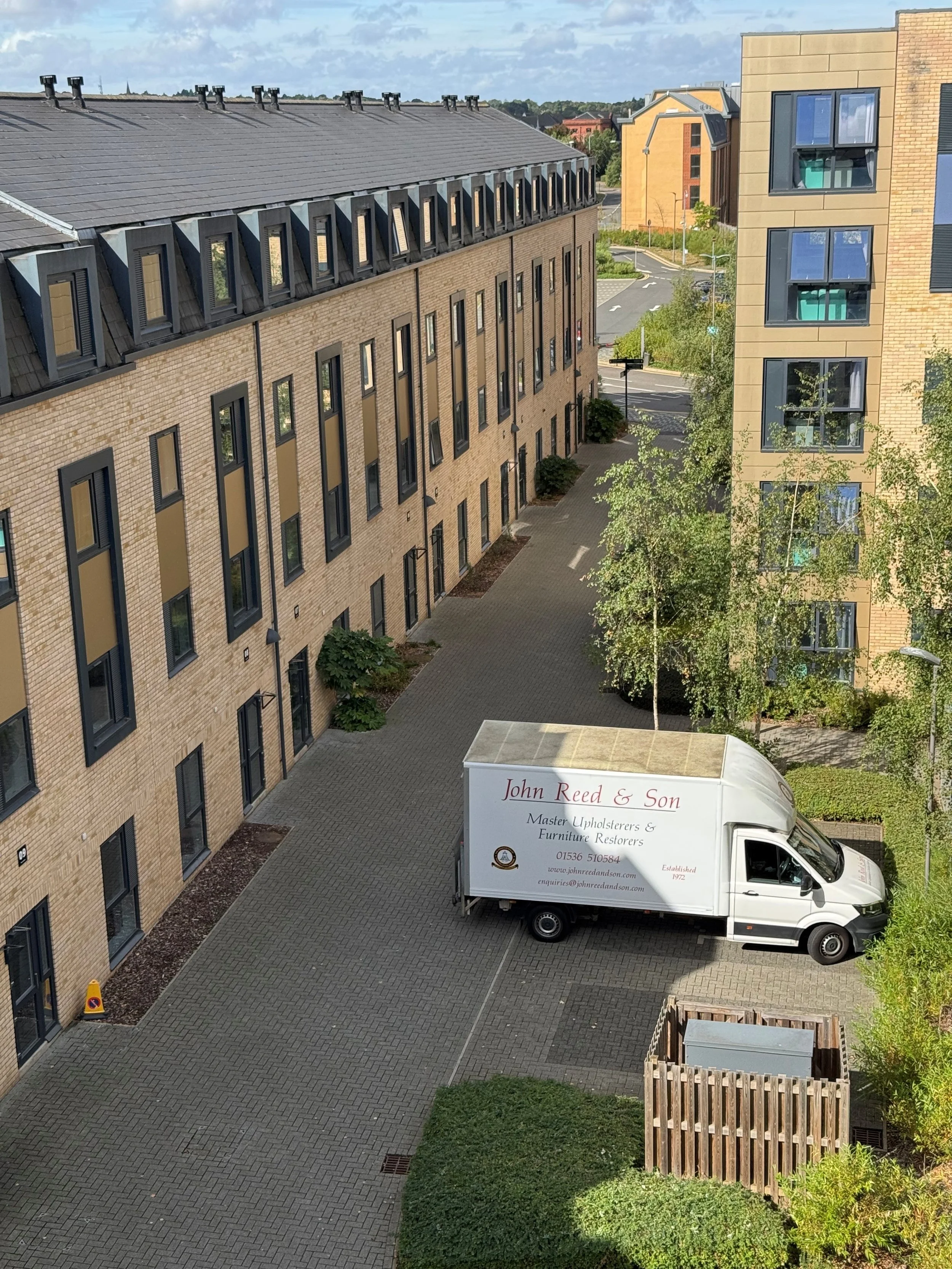 A cityscape featuring multi-story residential or commercial buildings with brick and modern facades, a paved courtyard area, small trees, and a white delivery truck with red and black text on it parked in the courtyard.