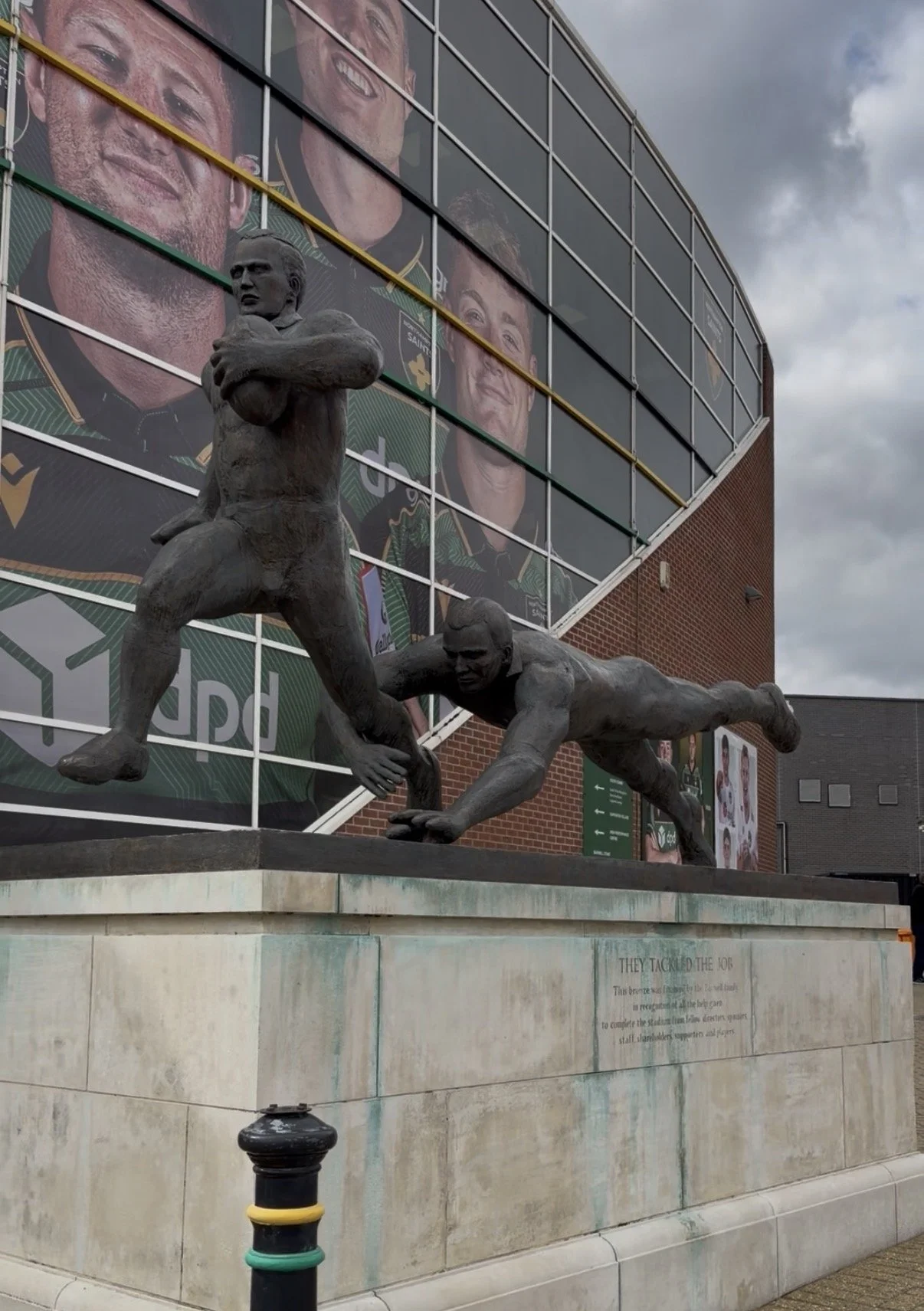 Scrumptuous bronze statues of two rugby players tackling each other, in front of a modern glass building with large portraits of rugby players on it.