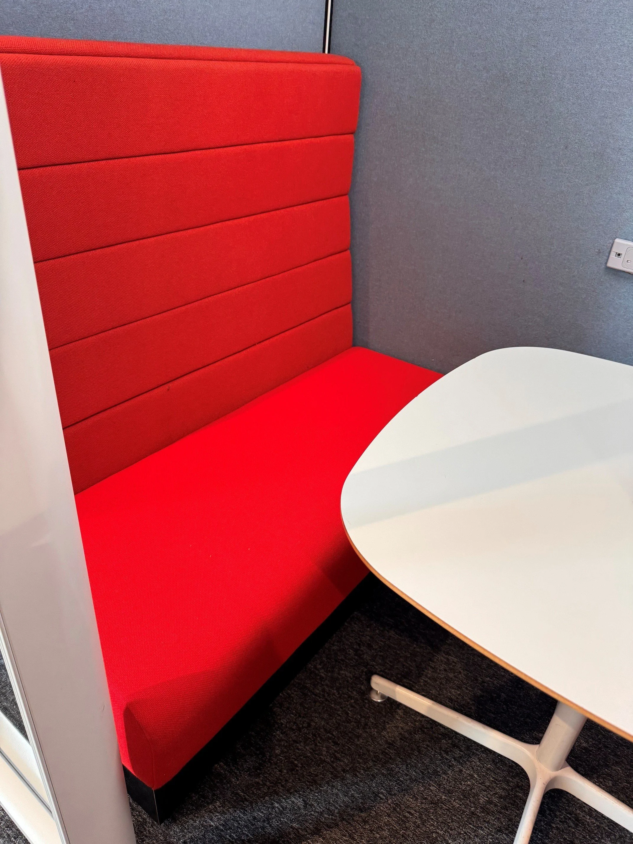 A red cushioned booth with a white table and a white chair in an office or lounge area, with gray carpeted flooring and a gray wall with a power outlet.