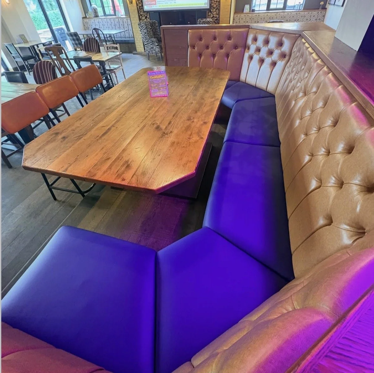 A wooden table surrounded by tan leather and dark purple cushioned seating in a restaurant or cafe, with a pink menu sign on the table and additional seating and decor in the background.