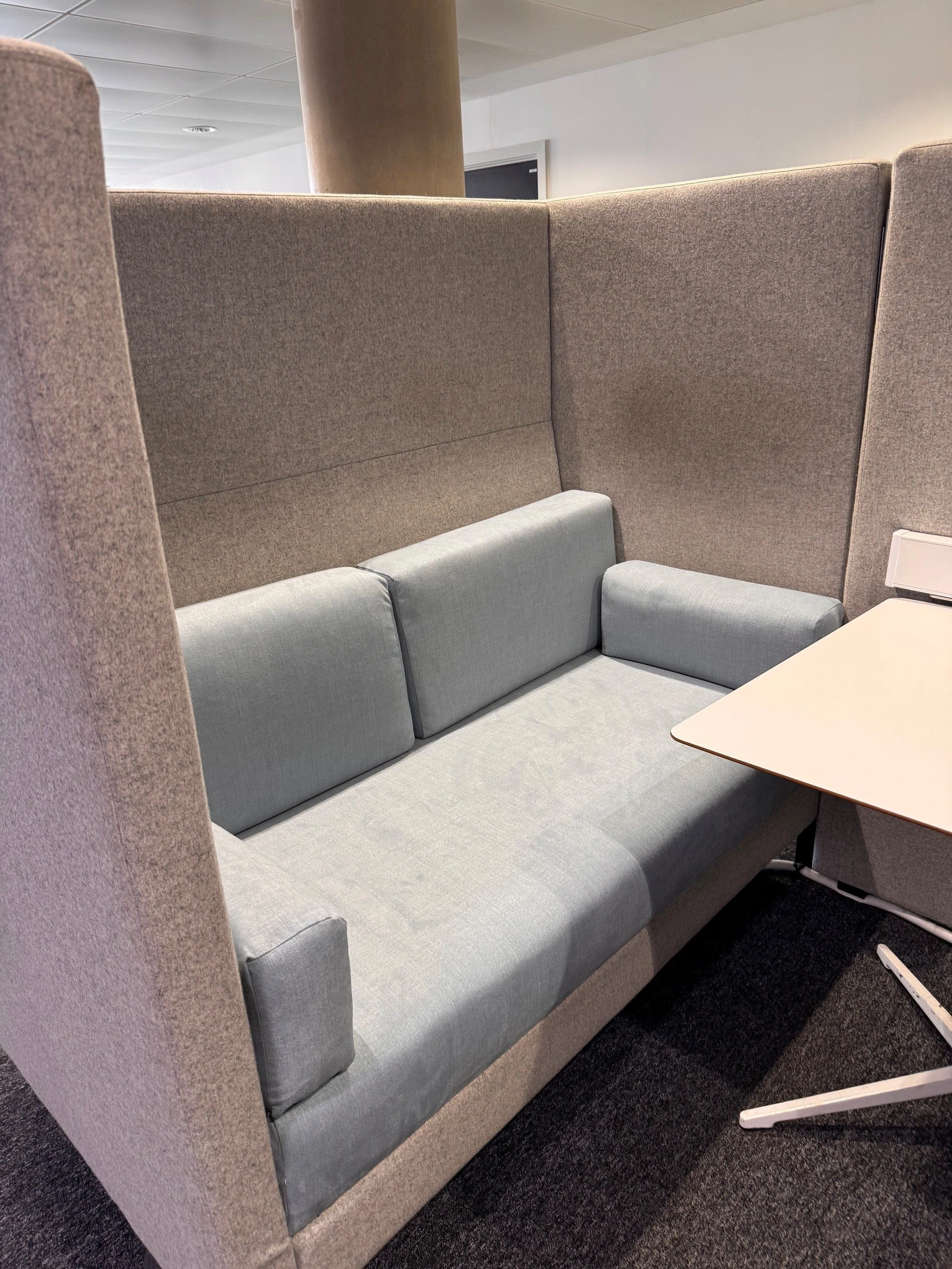 A modern office booth with light grey cushioned seats and high beige fabric walls.