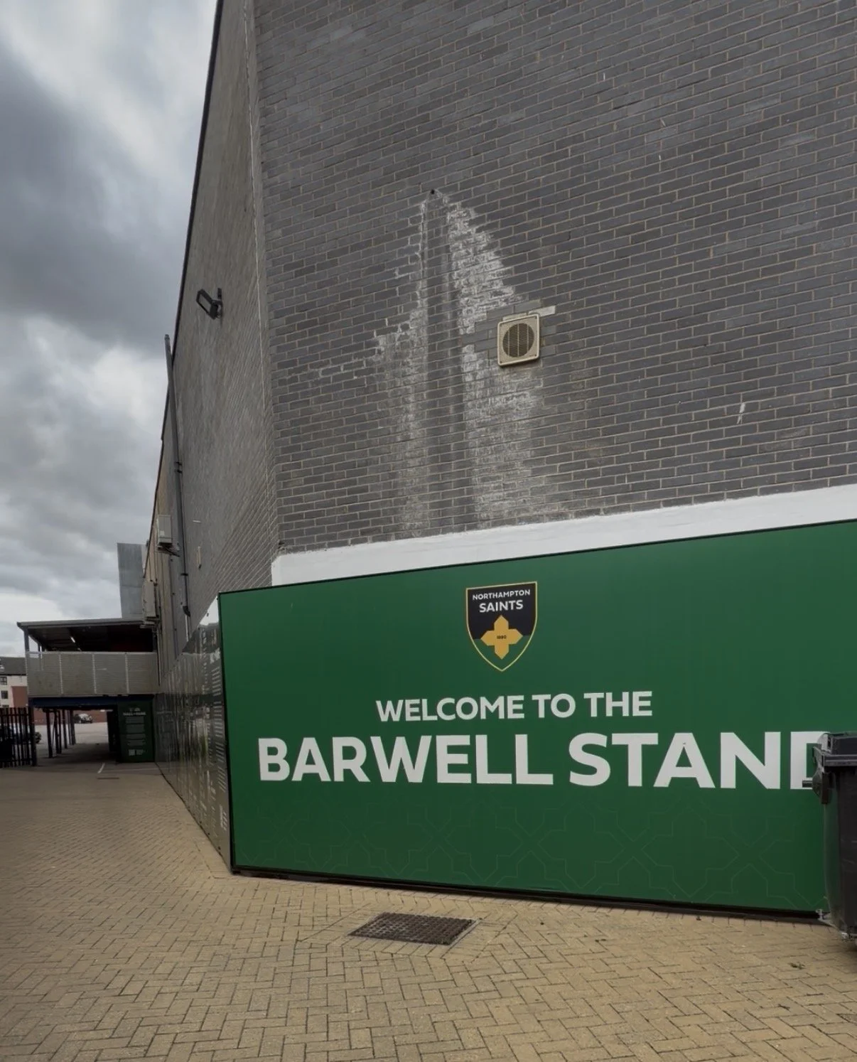 A large green sign reading "Welcome to the Barwell Stand" with a Northampton Saints logo above, situated next to a brick building with some white streaks and a vent, on a paved outdoor area under a cloudy sky.