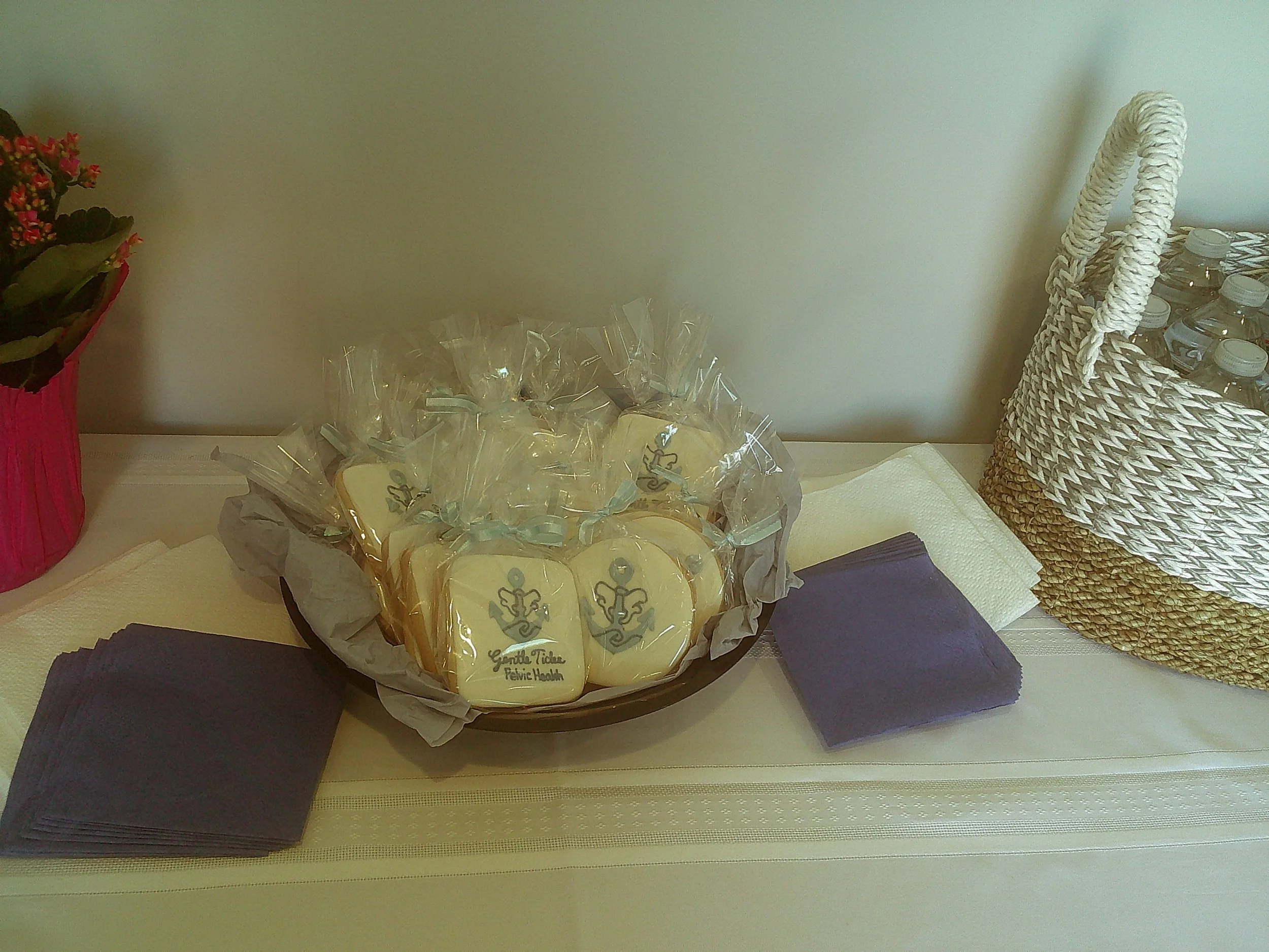 A basket of cookies with a label that says 'Gentle Tickle Felic Health,' next to a bouquet of pink and red flowers and a pile of purple napkins on a white table.