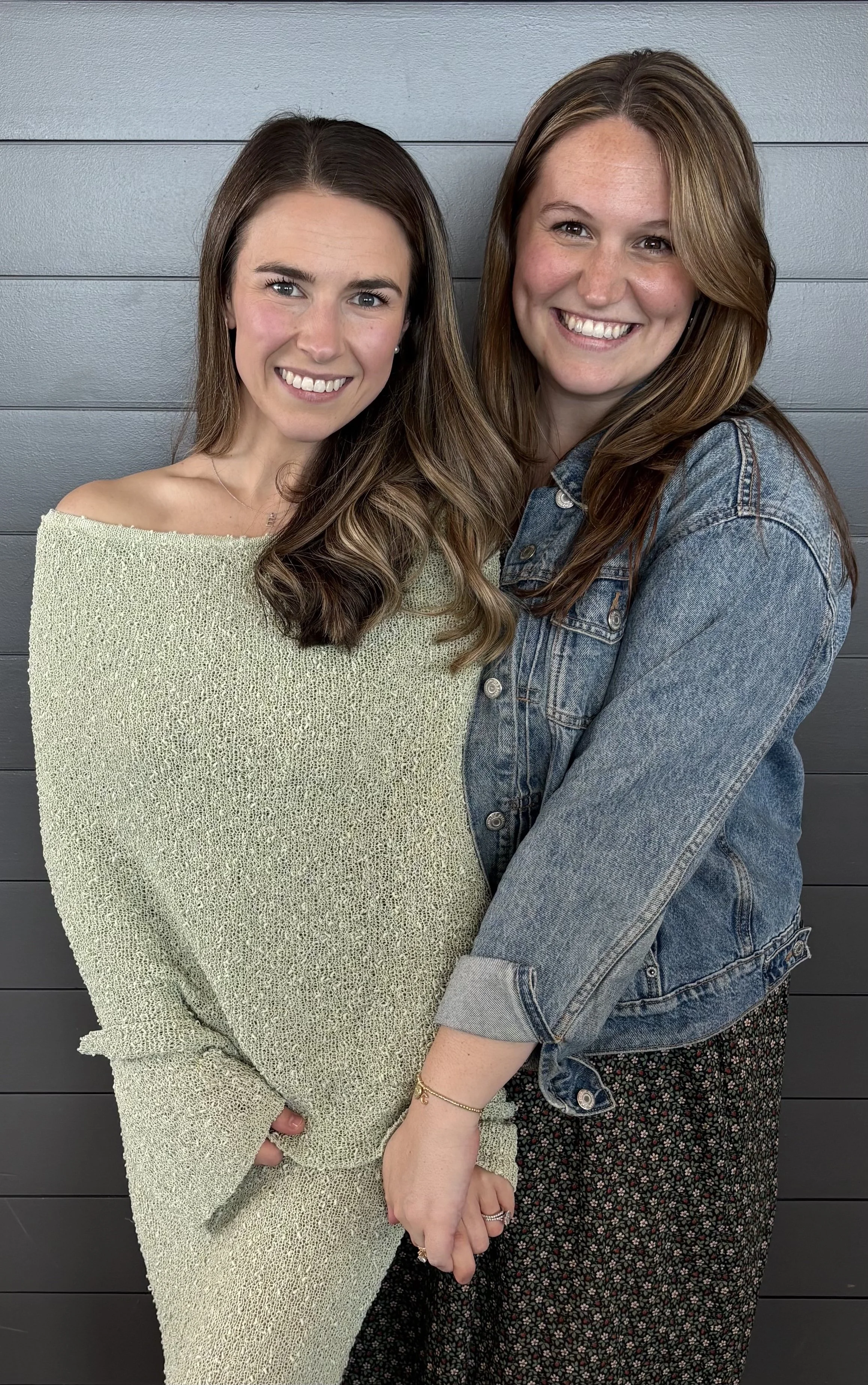Two women smiling and holding hands in front of a gray wall.