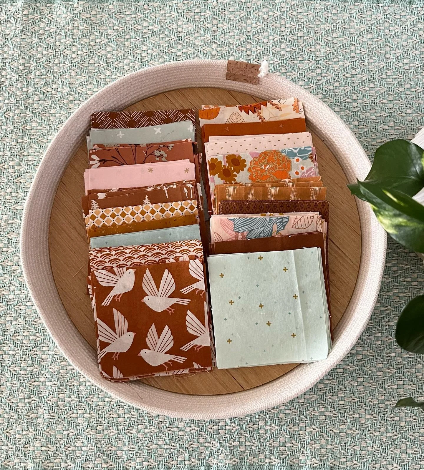A round woven basket with a wooden base, filled with various patterned paper napkins in earthy tones, placed on a textured woven mat with a green plant partially visible on the right.