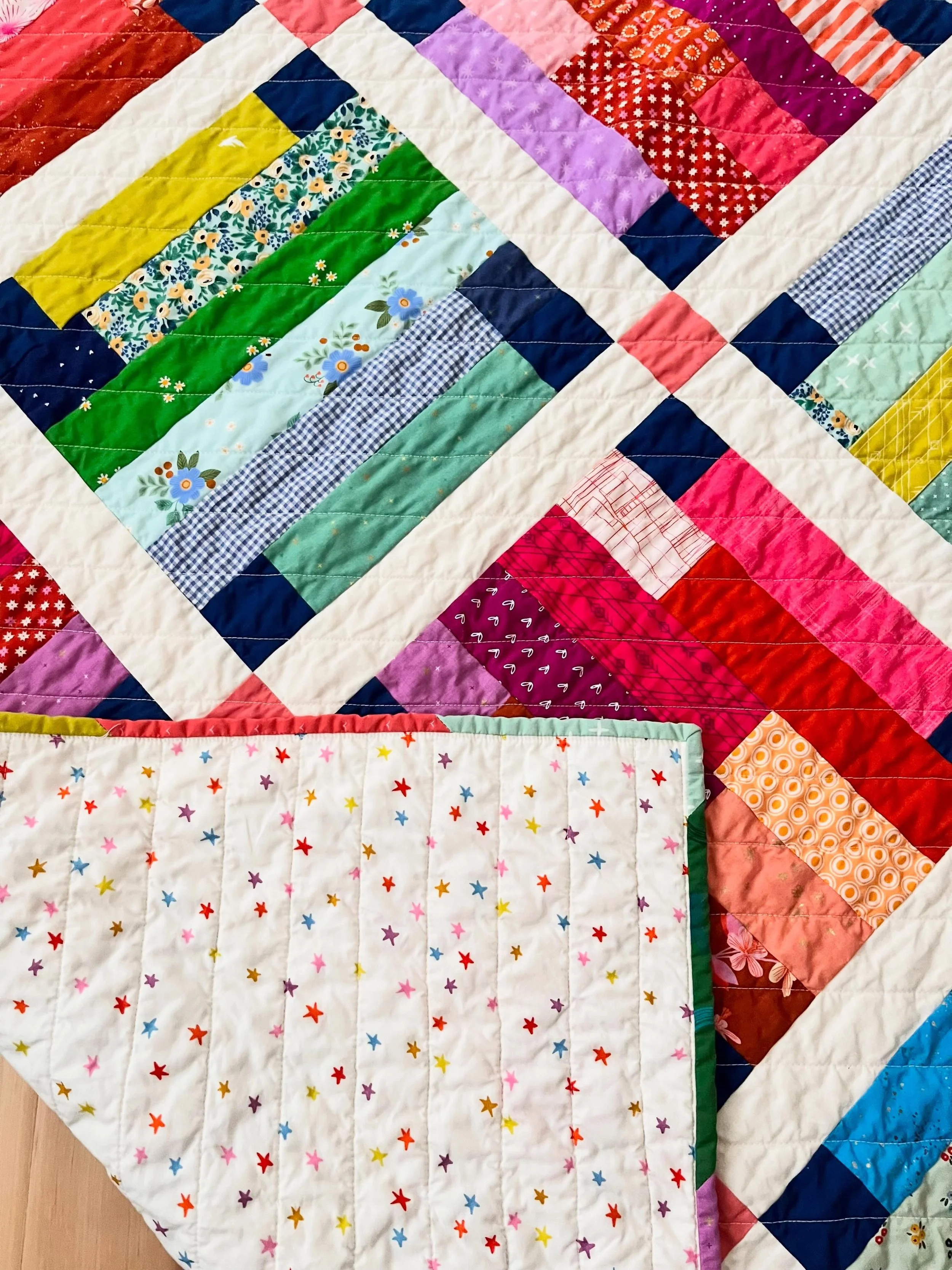 Close-up of a colorful patchwork quilt with various patterned fabric squares, including floral, checkered, and geometric designs, laid on a wooden floor.