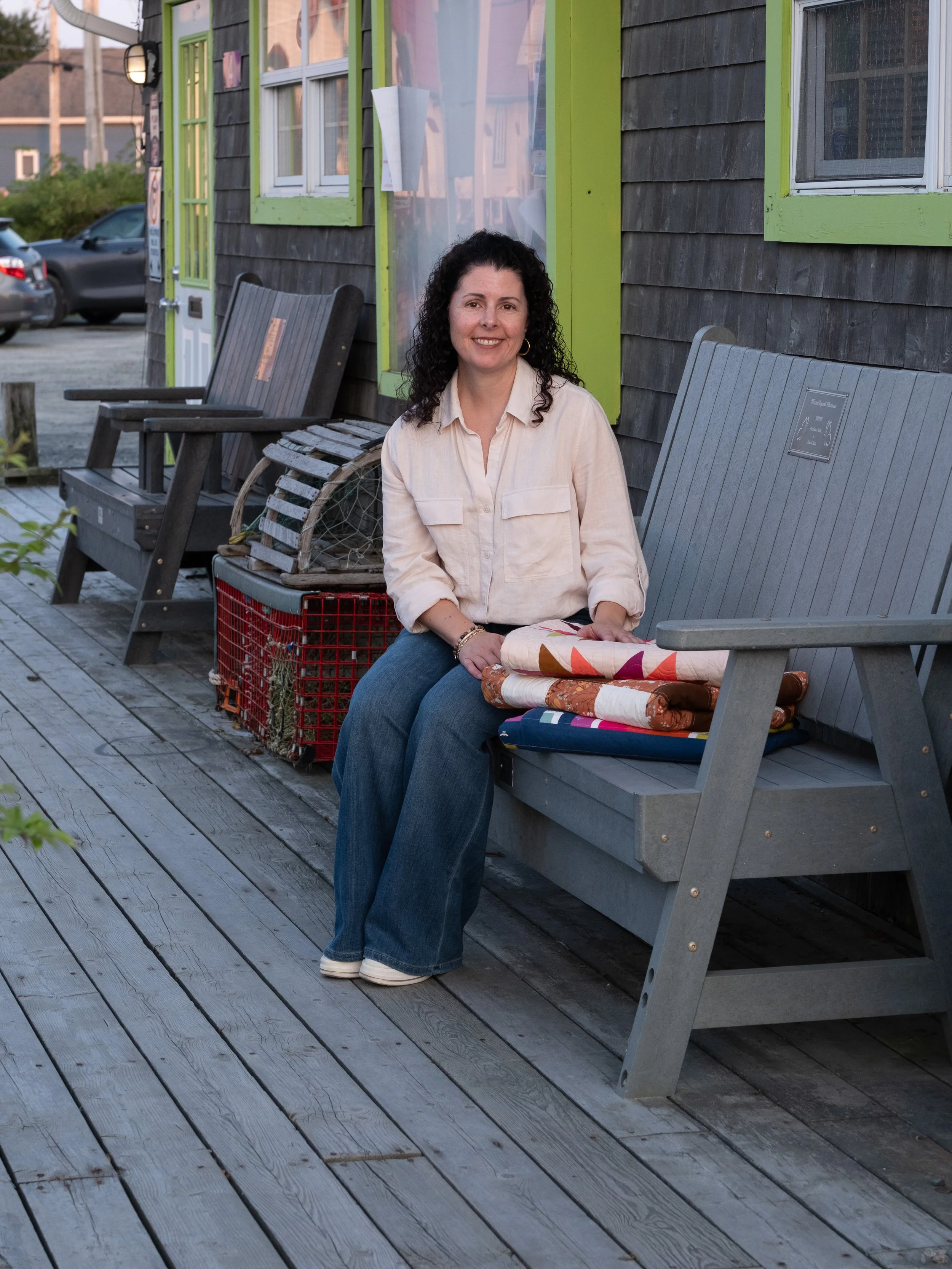 A woman with curly black hair, wearing a beige shirt and blue jeans, sitting on a gray wooden bench outside a building with a dark wooden siding and bright green window frames.