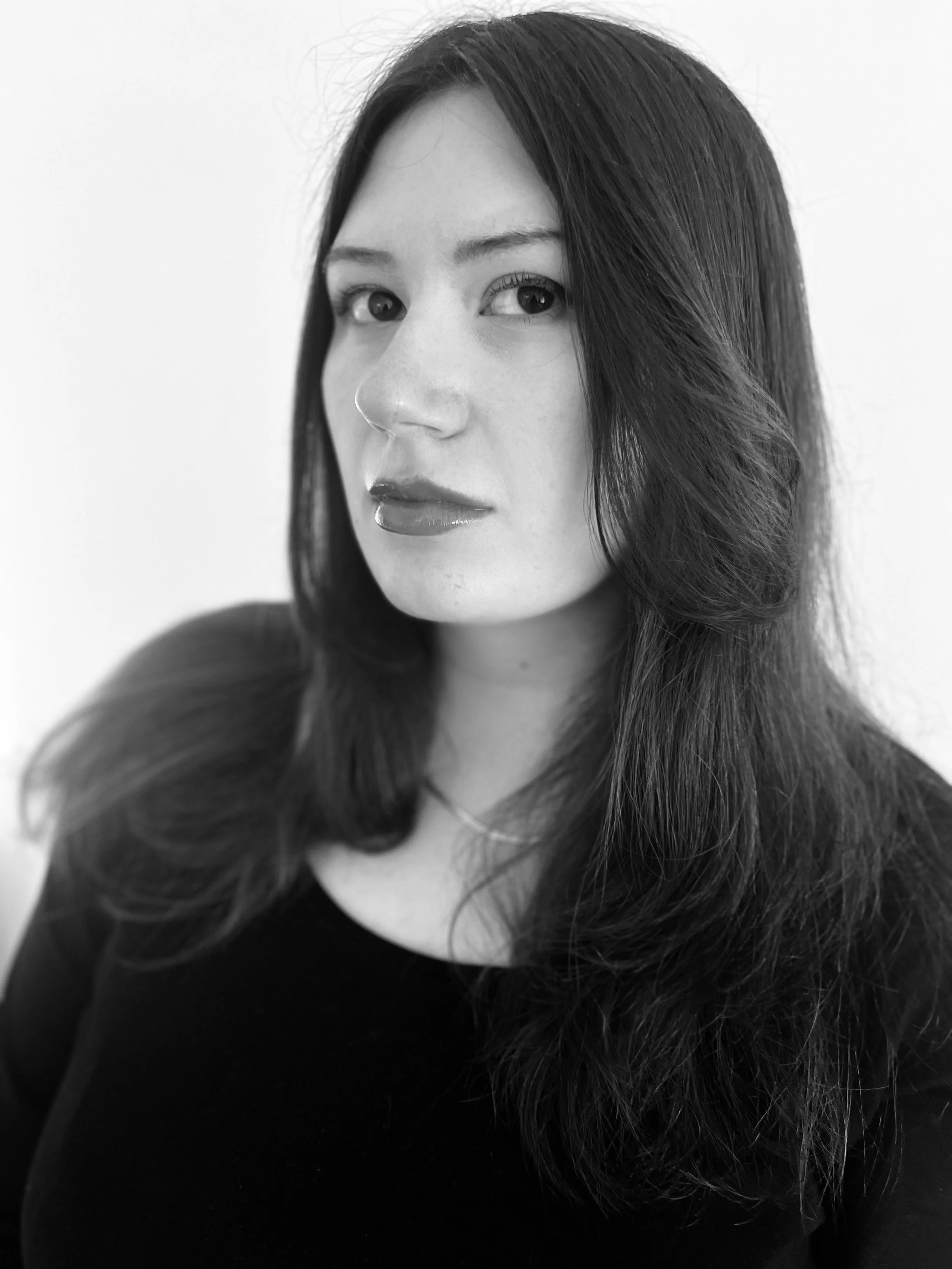 A black-and-white portrait of a woman with long, dark hair, wearing a dark top, looking over her shoulder.