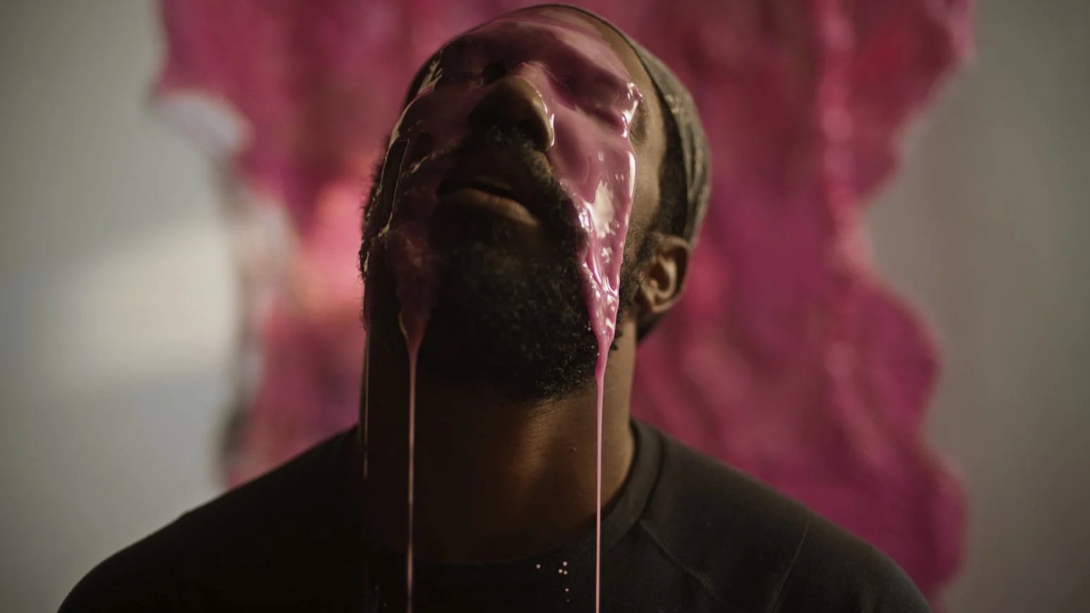A man with a beard and wearing a head covering has pink liquid dripping down his face against a pink abstract background.