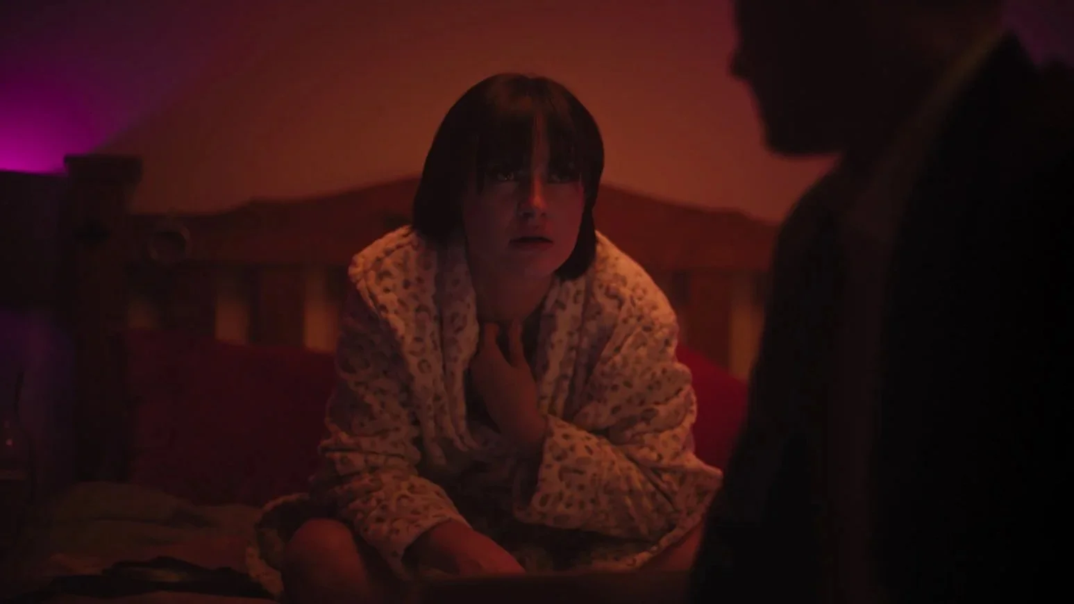 A woman with short dark hair sitting on a bed, wearing a fluffy patterned robe, with a distressed expression, holding her neck, illuminated by dimred lighting, in a bedroom setting.
