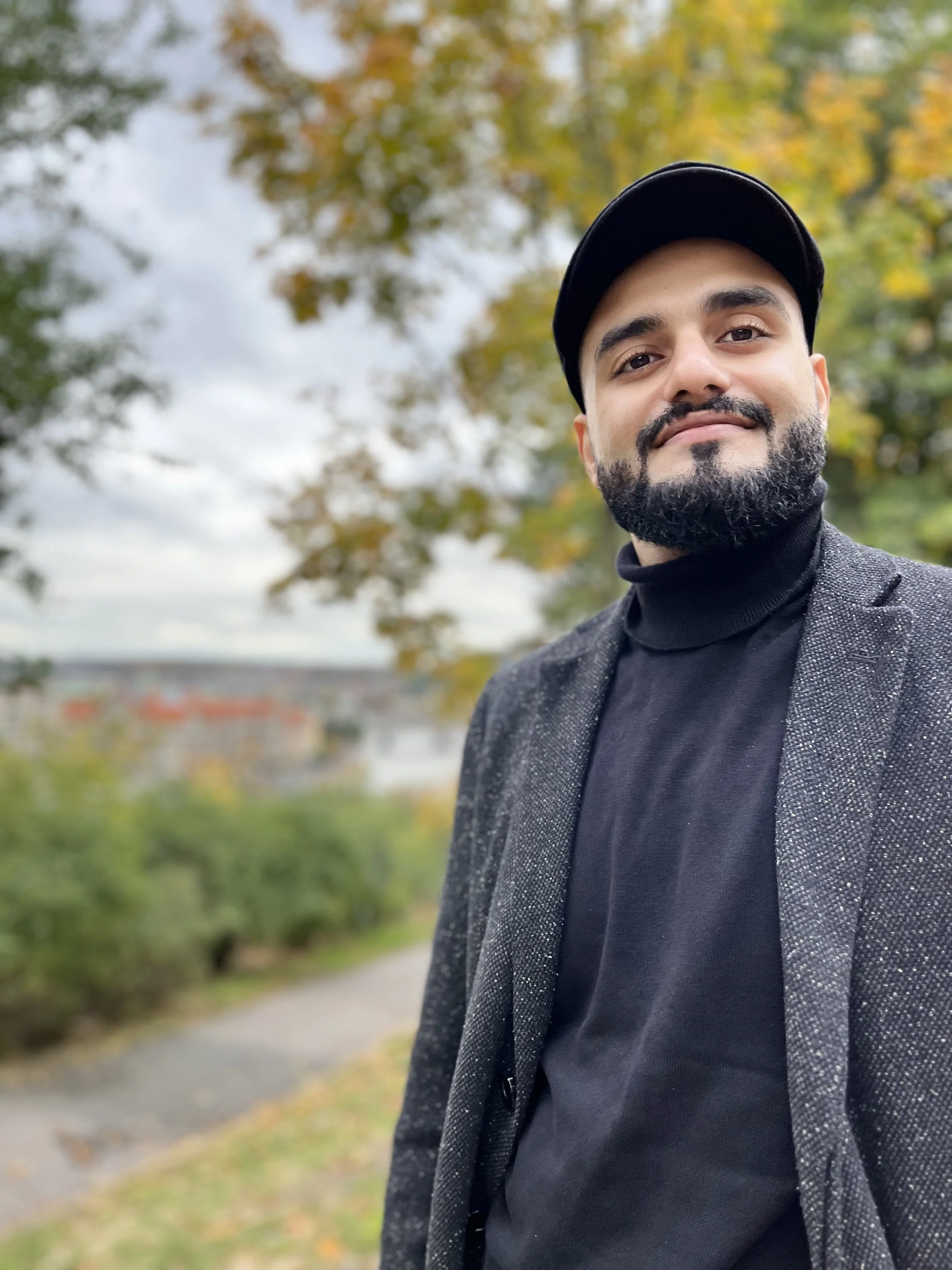 A man with a beard and mustache wearing a black baseball cap, black turtleneck sweater, and grey blazer taking a selfie outdoors with autumn-colored trees and cloudy sky in the background.