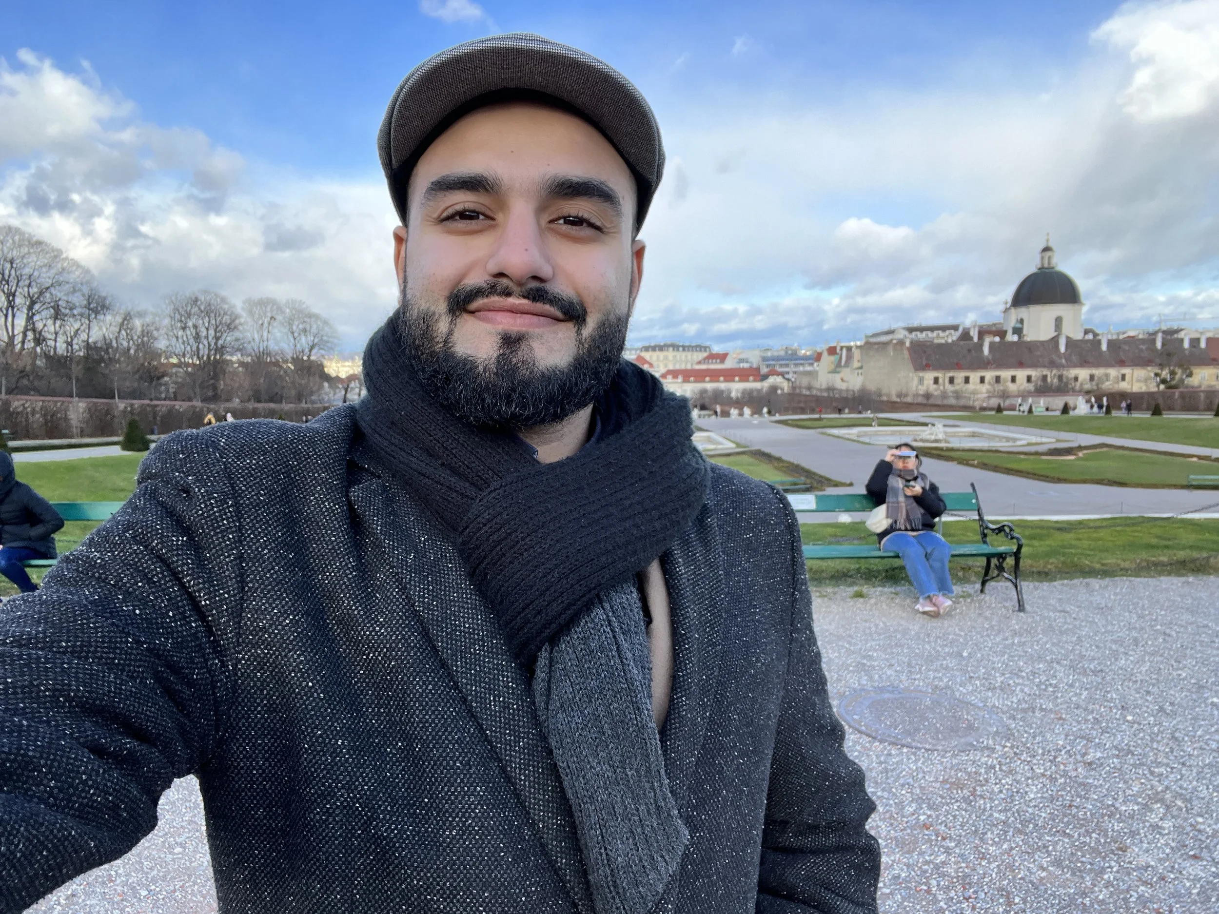 A close-up selfie of a man with a beard wearing a gray cap and a black scarf, standing in a park with benches, trees, and historic buildings in the background under a partly cloudy sky.