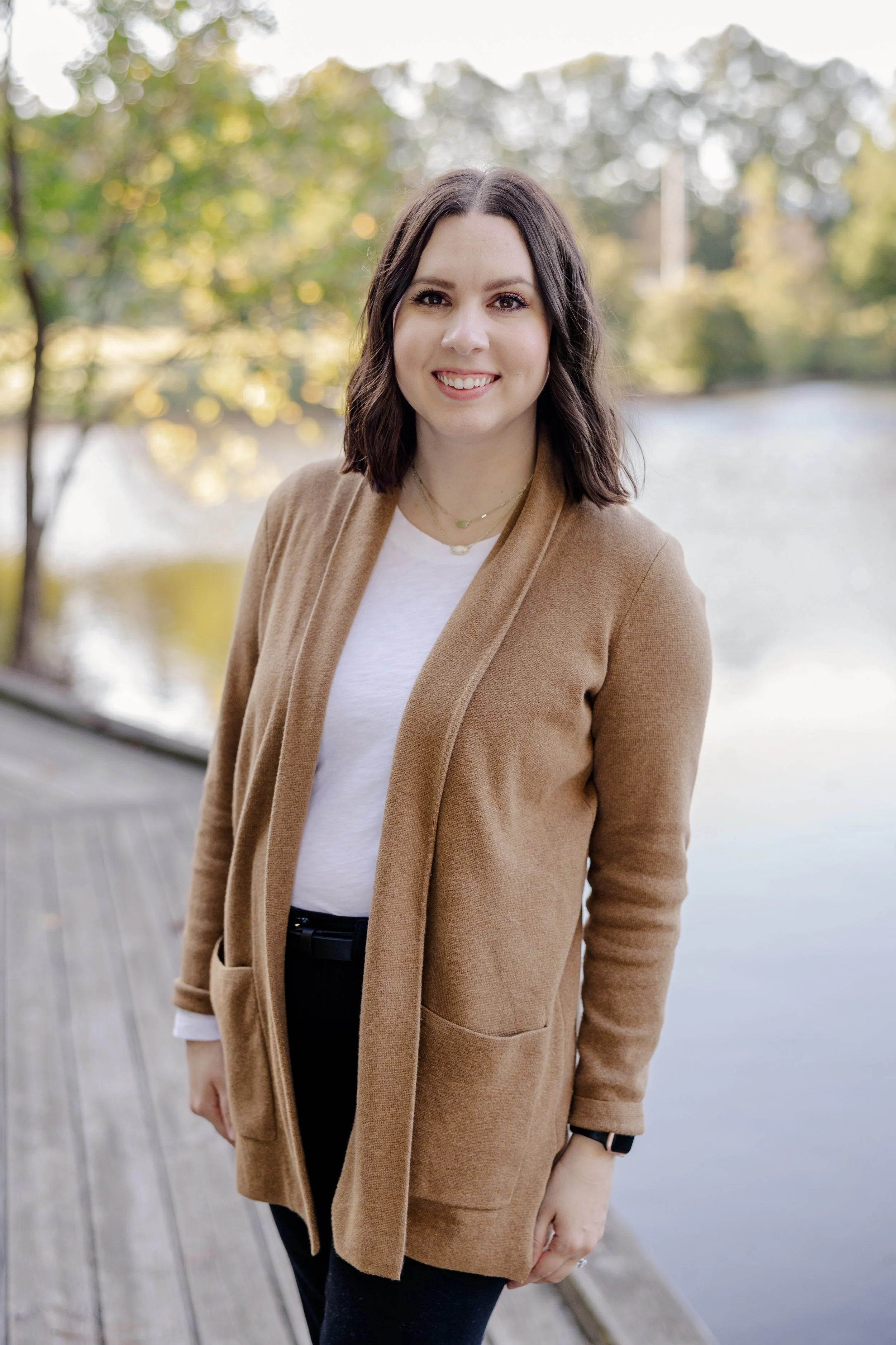 Young woman with shoulder-length dark hair smiling outdoors near a body of water, wearing a tan coat over a white shirt and black pants.