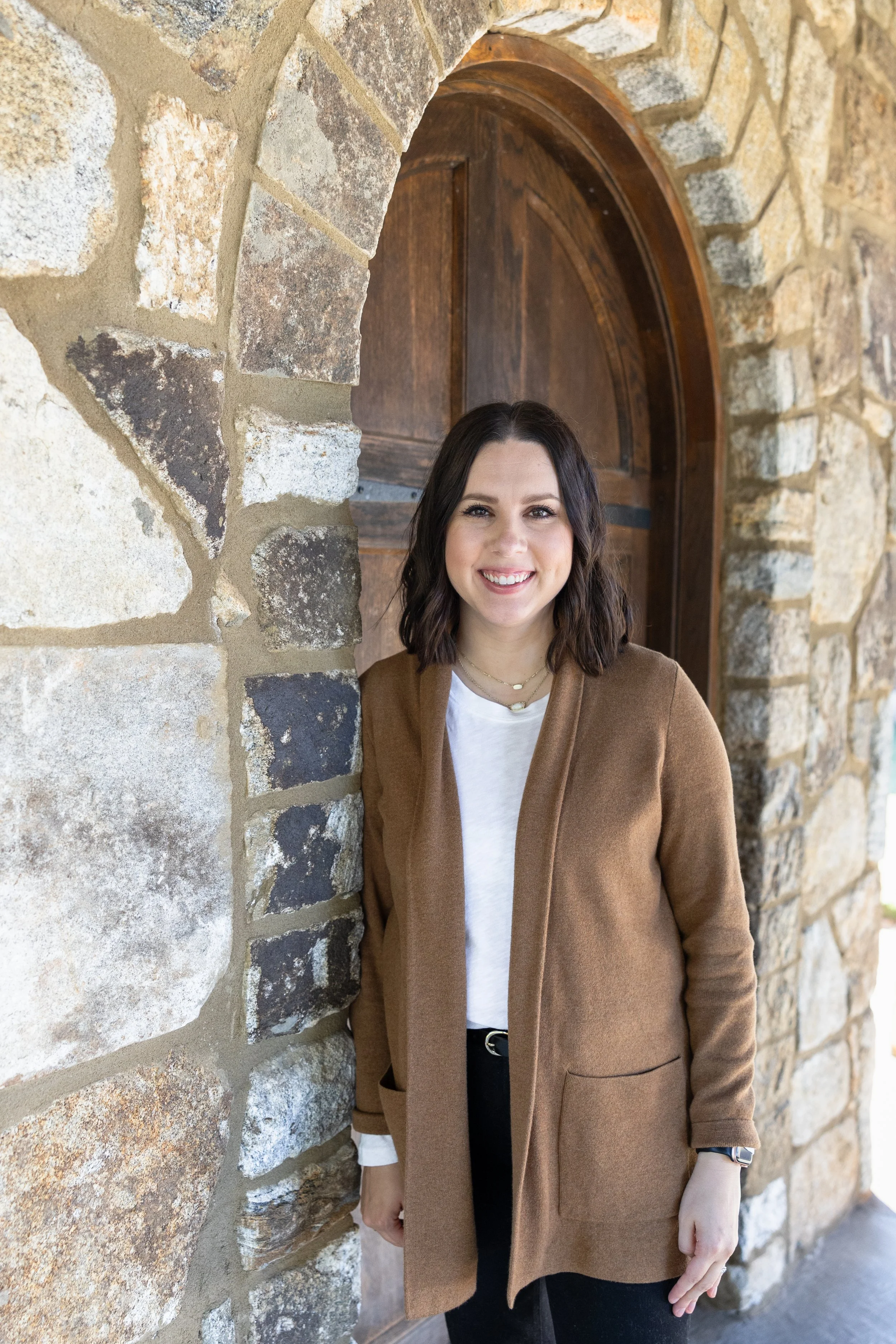 A smiling young woman with dark brown hair and light skin, wearing a brown coat over a white shirt, standing in front of a stone wall with a wooden arched door behind her.
