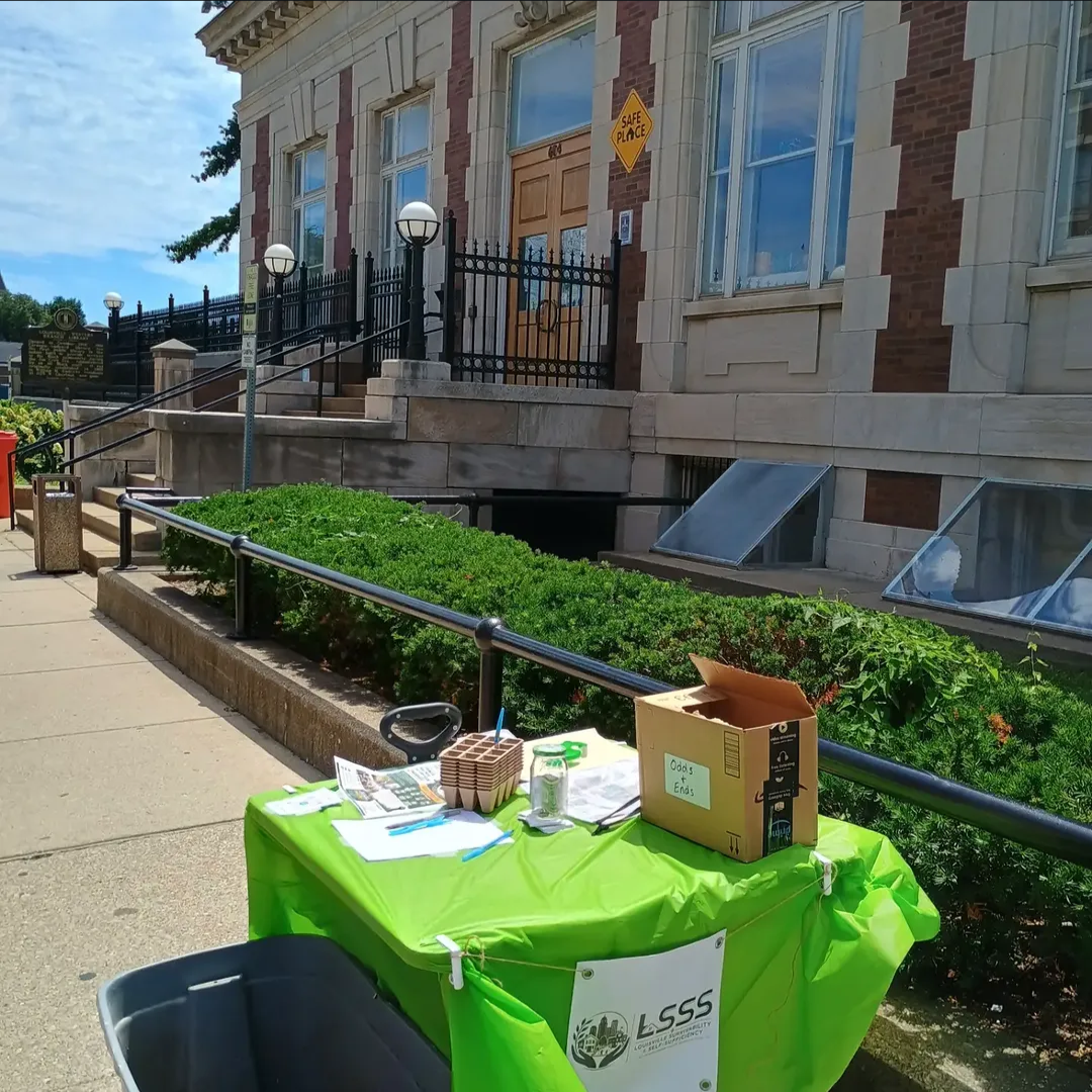 A street-level table covered with a bright green tablecloth, set up outdoors with various papers, a jar, a box labeled 'Odds & Ends,' and other small items. The table is next to a sidewalk in front of a historic-looking building with stone and brick 