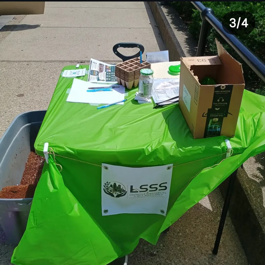 A green-covered table set up outdoors with various items including a box, a jar, a clipboard, printed papers, blue pens, a stack of paper plates, and a carabiner. There is a sign on the front of the table that reads 'LSS Louisville Survivability & Se
