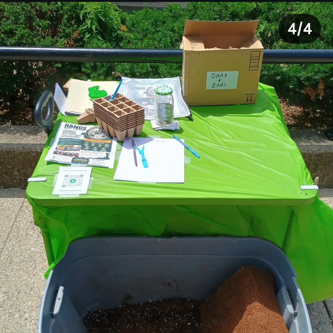 A gardening setup on a bright green table with a cardboard box, gardening tools, seed starting trays, a jar, a flyer, and a notebook, with a large container of soil in front, outdoors with bushes in the background.