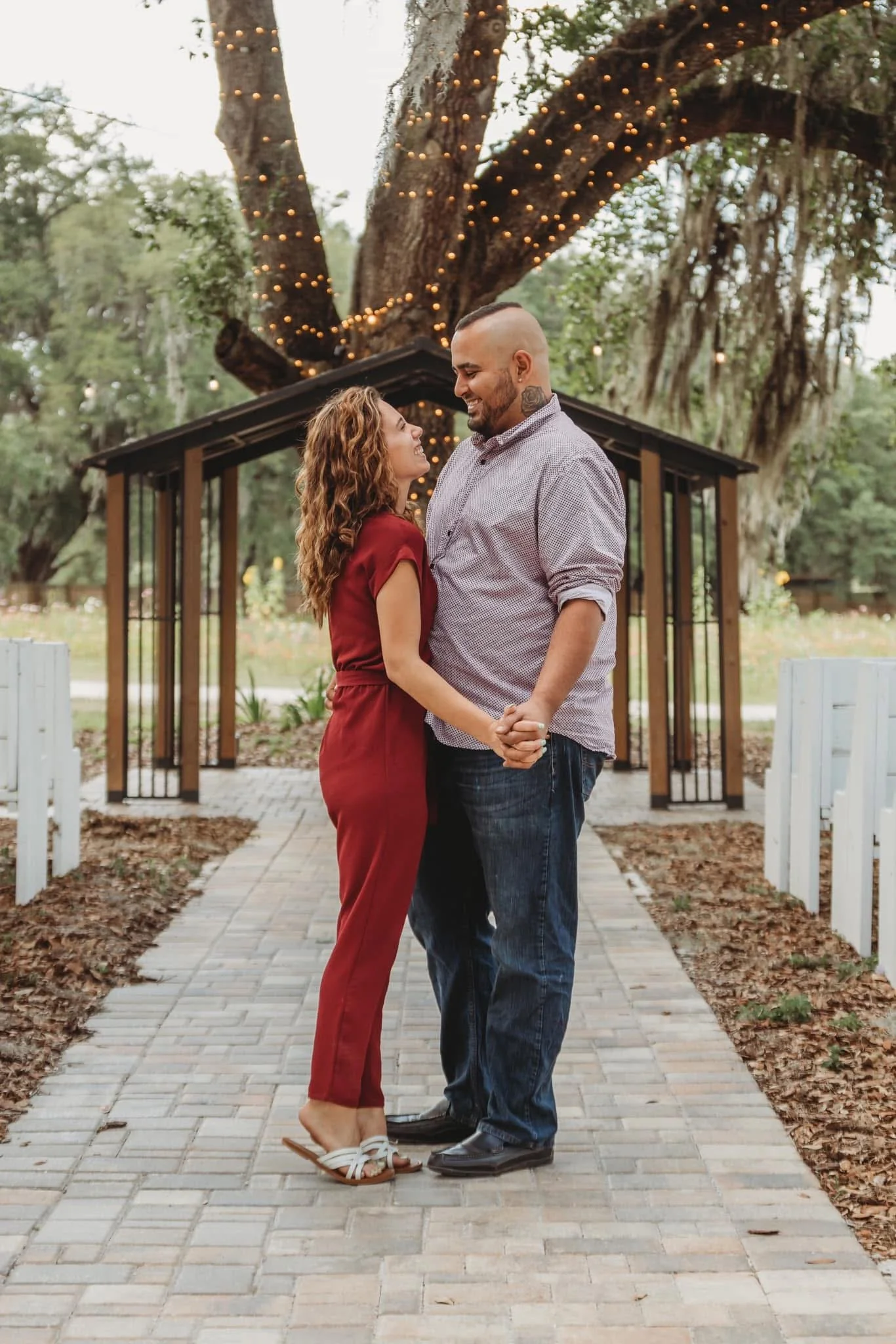 A couple stands close together outdoors, holding hands and smiling at each other, under a large tree decorated with string lights, with a small building and trees in the background.