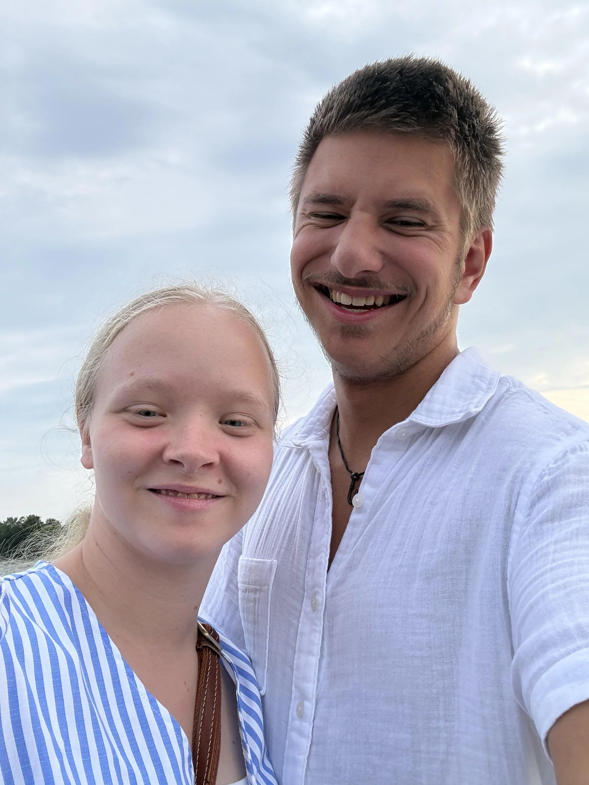A smiling man with short dark hair wearing a white shirt standing outdoors with a woman who has blonde hair and light skin in front of him. The sky is visible in the background.