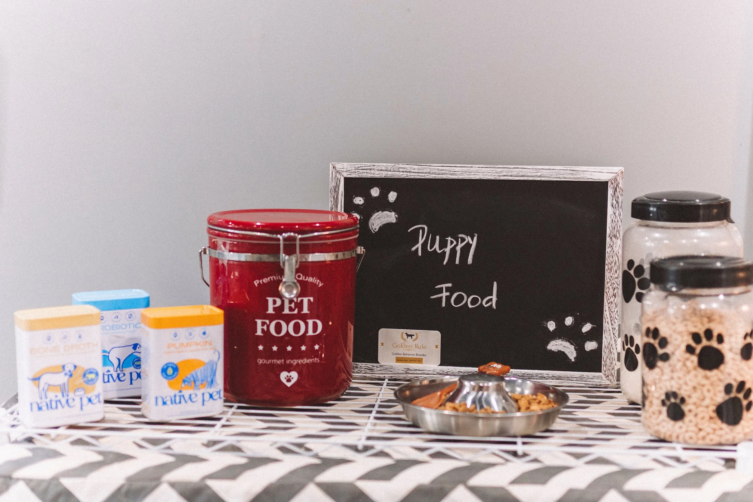 Collection of pet food containers and treats on a table, including a red pet food canister, a row of small containers with dog treats, and a blackboard sign that says "Puppy Food" with paw print illustrations.
