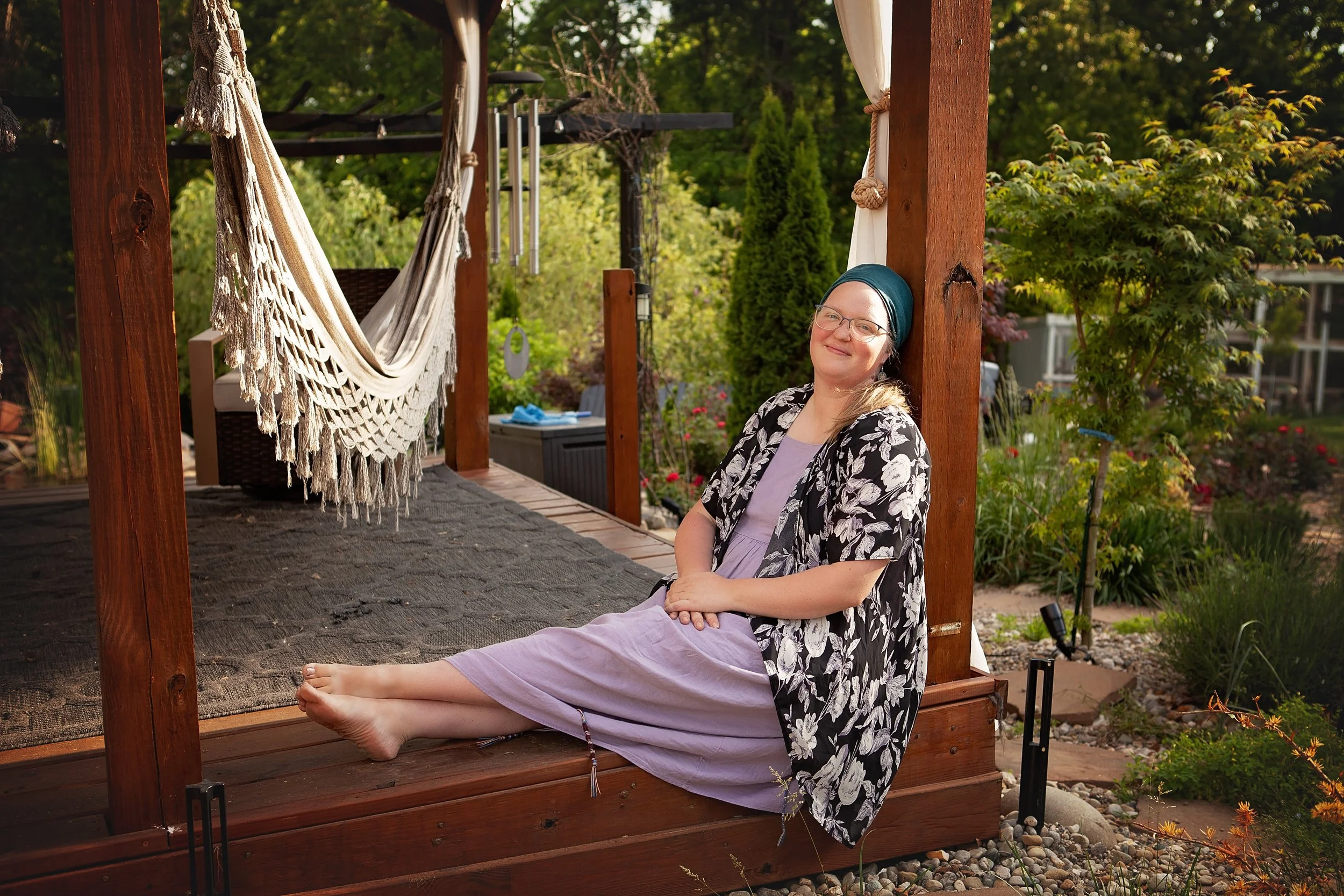 A woman with glasses and a headscarf sitting on a wooden porch swing, smiling, surrounded by greenery and garden plants.