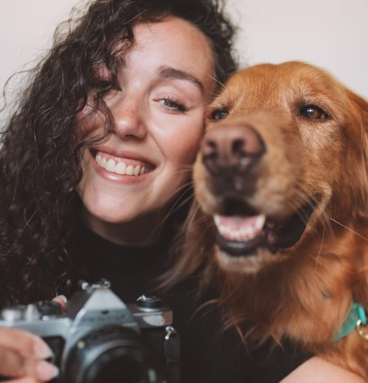A woman with dark curly hair smiling next to a golden retriever dog. The woman is holding a camera. The dog is happy with its mouth open. Both appear close and joyful.