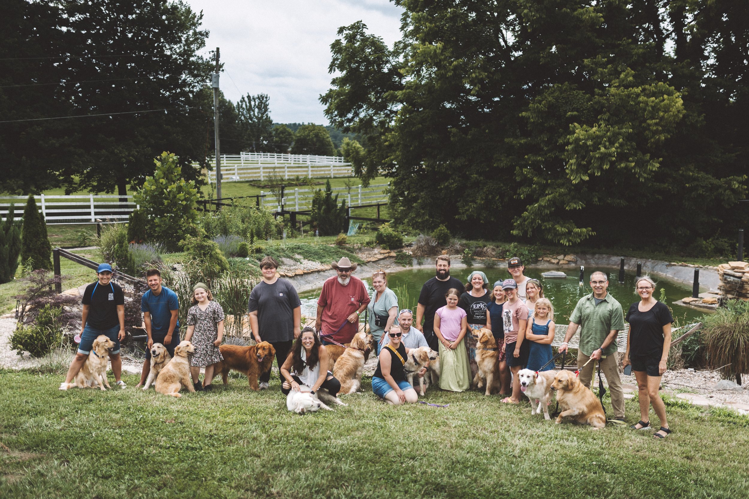 A large group of people and dogs posing together outdoors in a garden with greenery and trees, a pond, and a white fence in the background.