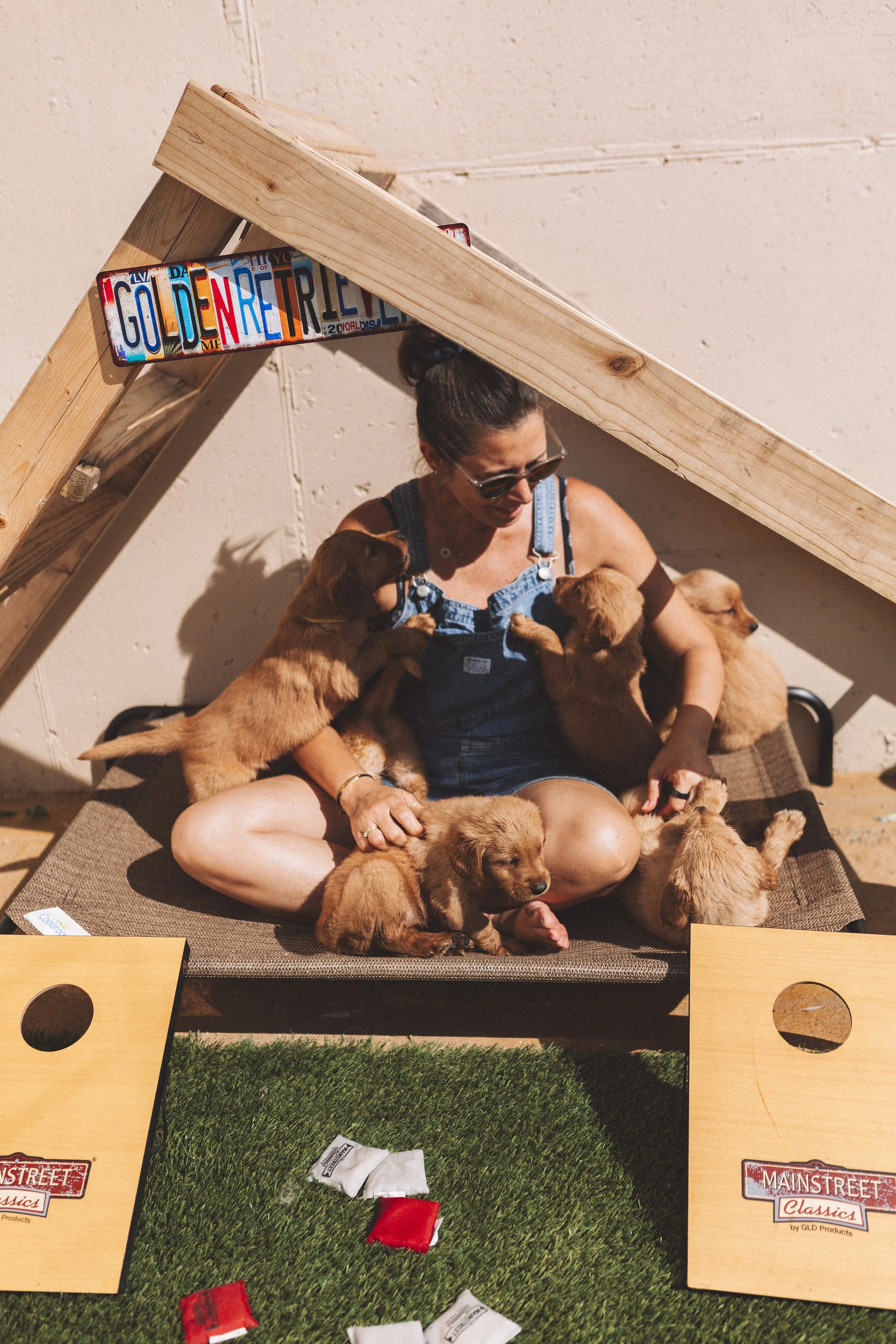 A woman sitting on a carpet playing a tabletop game with puppies around her. The game features a small green artificial grass area with red and white objects, and wooden rails with 'Main Street Classics' branding.