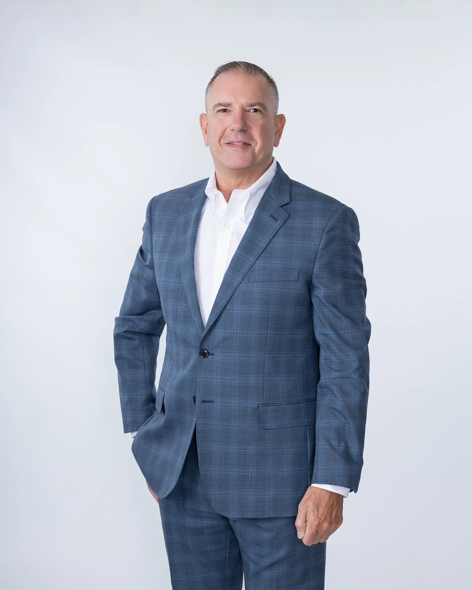 A middle-aged man with short gray hair wearing a white dress shirt and a blue checkered suit, standing against a plain white background.