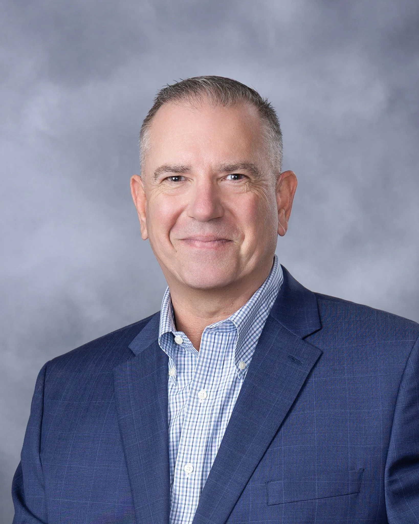 Headshot of a smiling middle-aged man with short gray hair, wearing a dark blue checked suit jacket and a light blue polo shirt, set against a gray background.