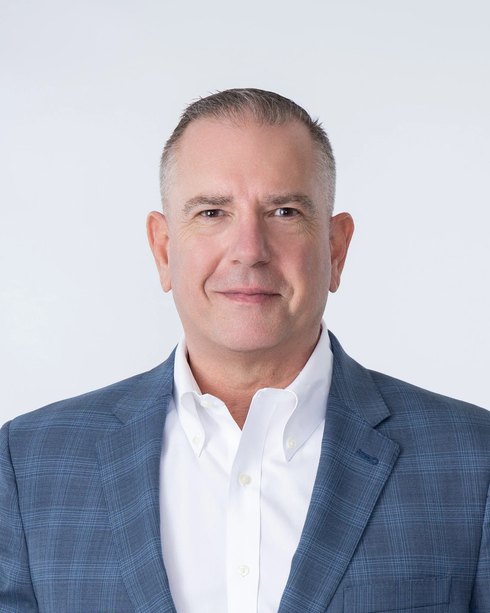 Close-up image of a middle-aged man wearing a blue plaid suit and white shirt, smiling gently against a plain light gray background.