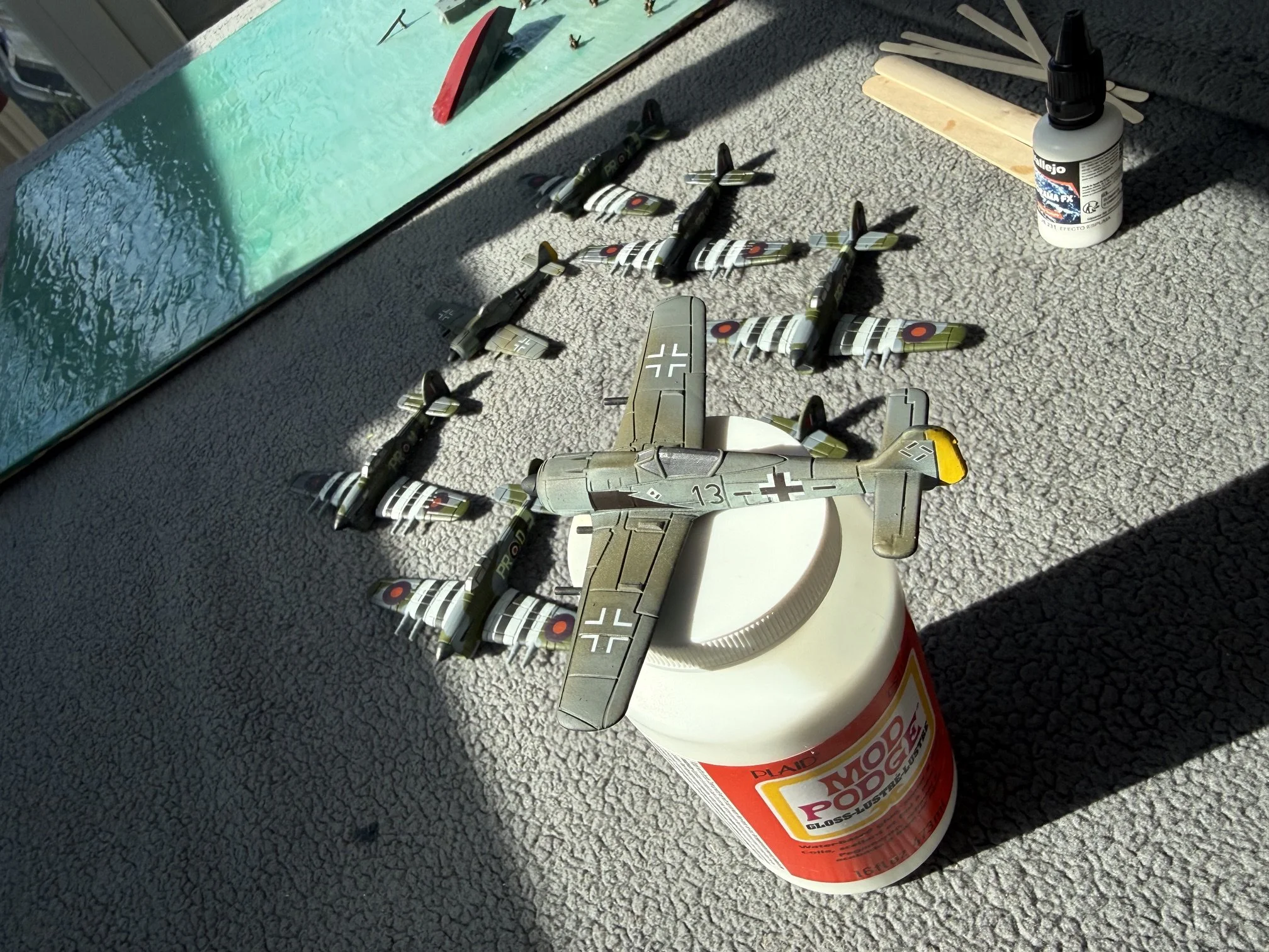 Model airplanes resembling World War II aircrafts are arranged on a container of glue on a textured gray surface. The scene is lit by bright sunlight, casting shadows.